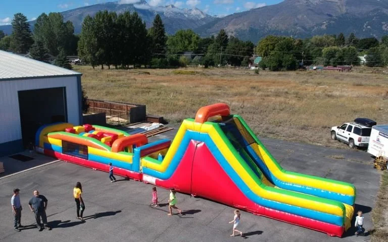 Colorful inflatable slide and obstacle course set up outdoors on a paved area, with a few children playing and adults nearby, against a backdrop of trees, mountains, and a blue sky.