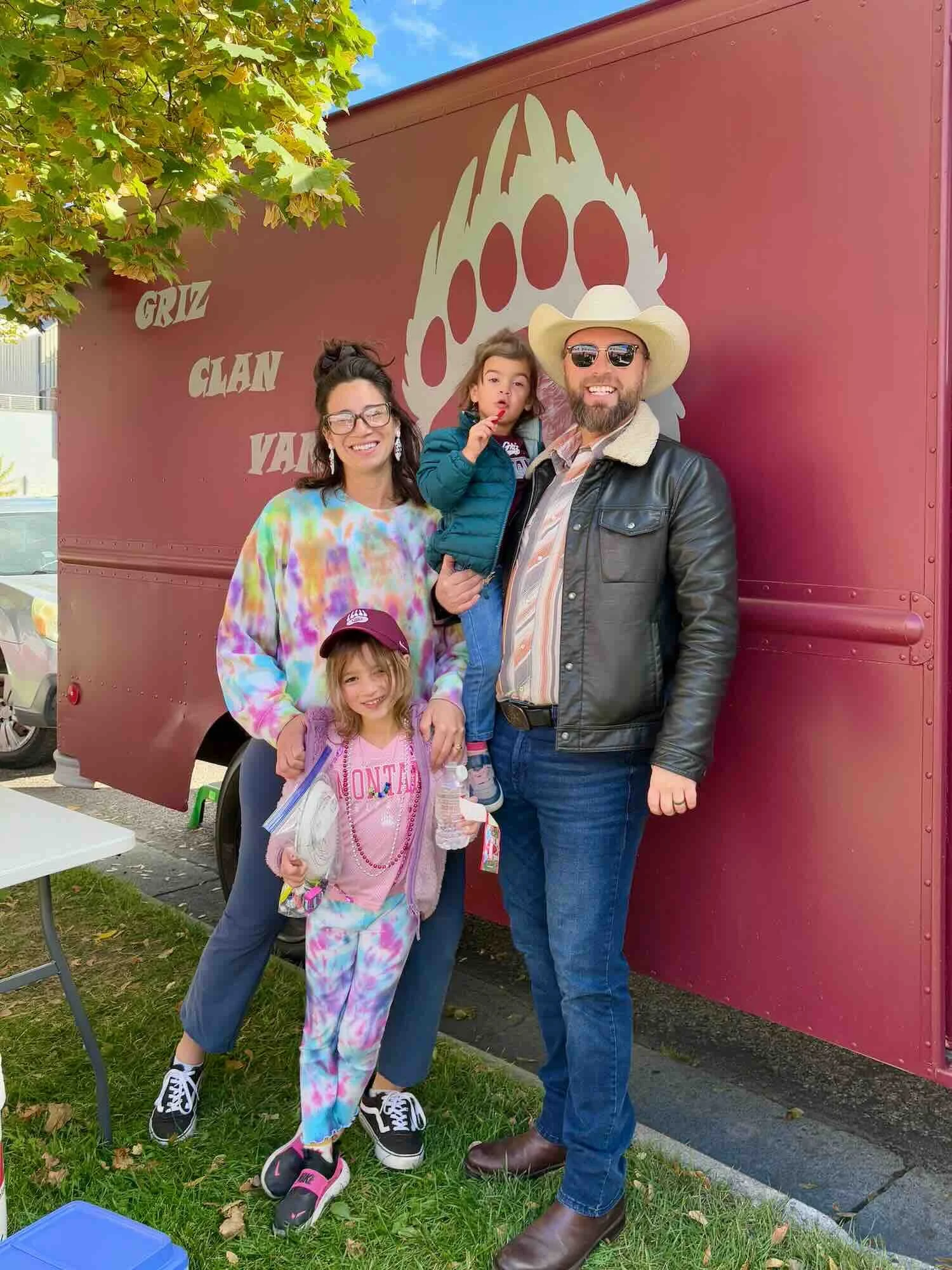A family of four, including a man with a cowboy hat, sunglasses, and a leather jacket, a woman with glasses and colorful shirt, a young girl in tie-dye clothing, and a young girl in a pink shirt and cap, standing outdoors near a maroon trailer with a bear paw print and the words 'CRUZ CLAN YARD' painted on it, surrounded by green grass and trees.