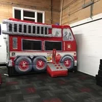 A red and gray fire truck-shaped playhouse for children, featuring a ladder, windows, and a slide on a black rubber mat floor.