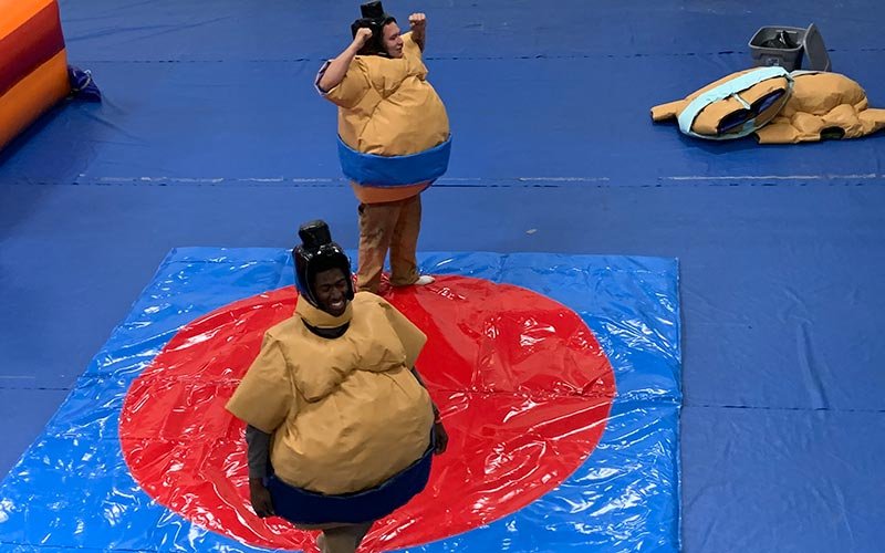 Two people dressed in inflatable sumo wrestling suits, smiling and standing on a red and blue mat inside an indoor facility.