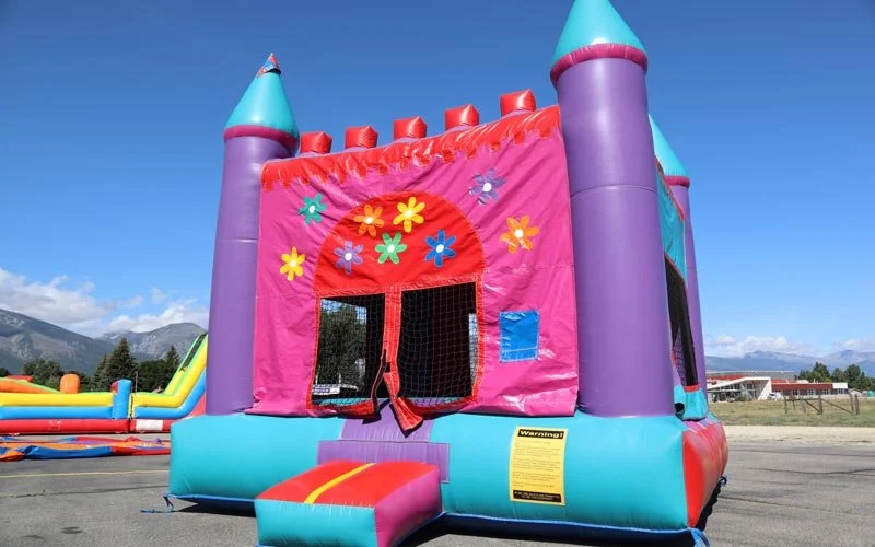 Colorful inflatable bounce house castle with flower decorations and a small red slide, set outdoors on a sunny day.