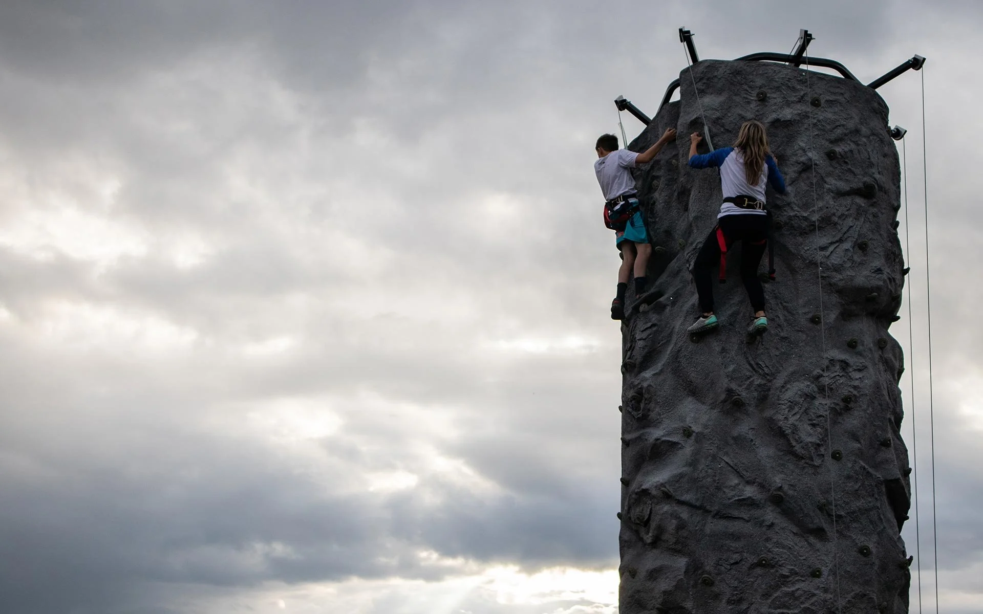 Two people rock climbing on an outdoor artificial climbing wall under a cloudy sky.