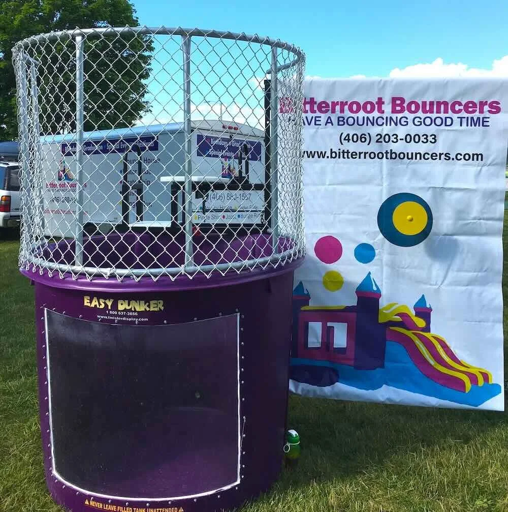 A bounce house with a circular, enclosed mesh top, branded as 'Easy Punker,' situated on a grassy area next to a colorful banner advertising 'Bitterroot Bouncers,' a bounce house rental company.