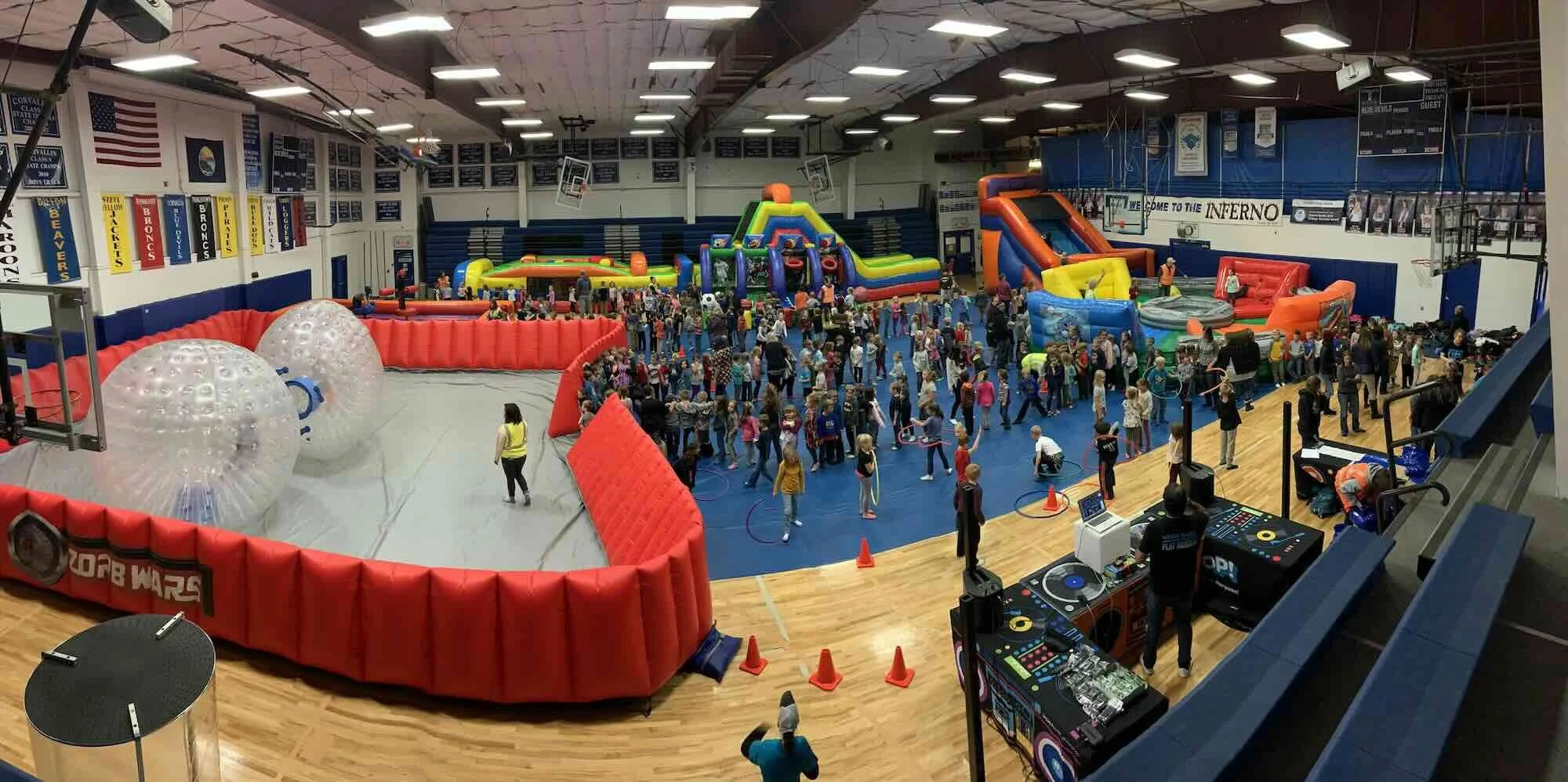 An indoor trampoline park filled with children and adults. There are various inflatable bounce houses, spherical bumper ball arenas, and a DJ setup near the foreground. The venue has banners and flags hanging from the walls, and a scoreboard visible on the back wall.