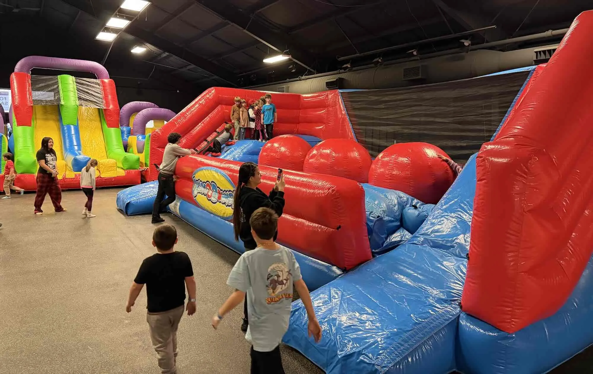 Indoor bounce house with multiple inflatable structures, children playing, and adults supervising. Brightly colored inflatables include a slide, climbing areas, and large balls, set in a spacious room with a dark ceiling.