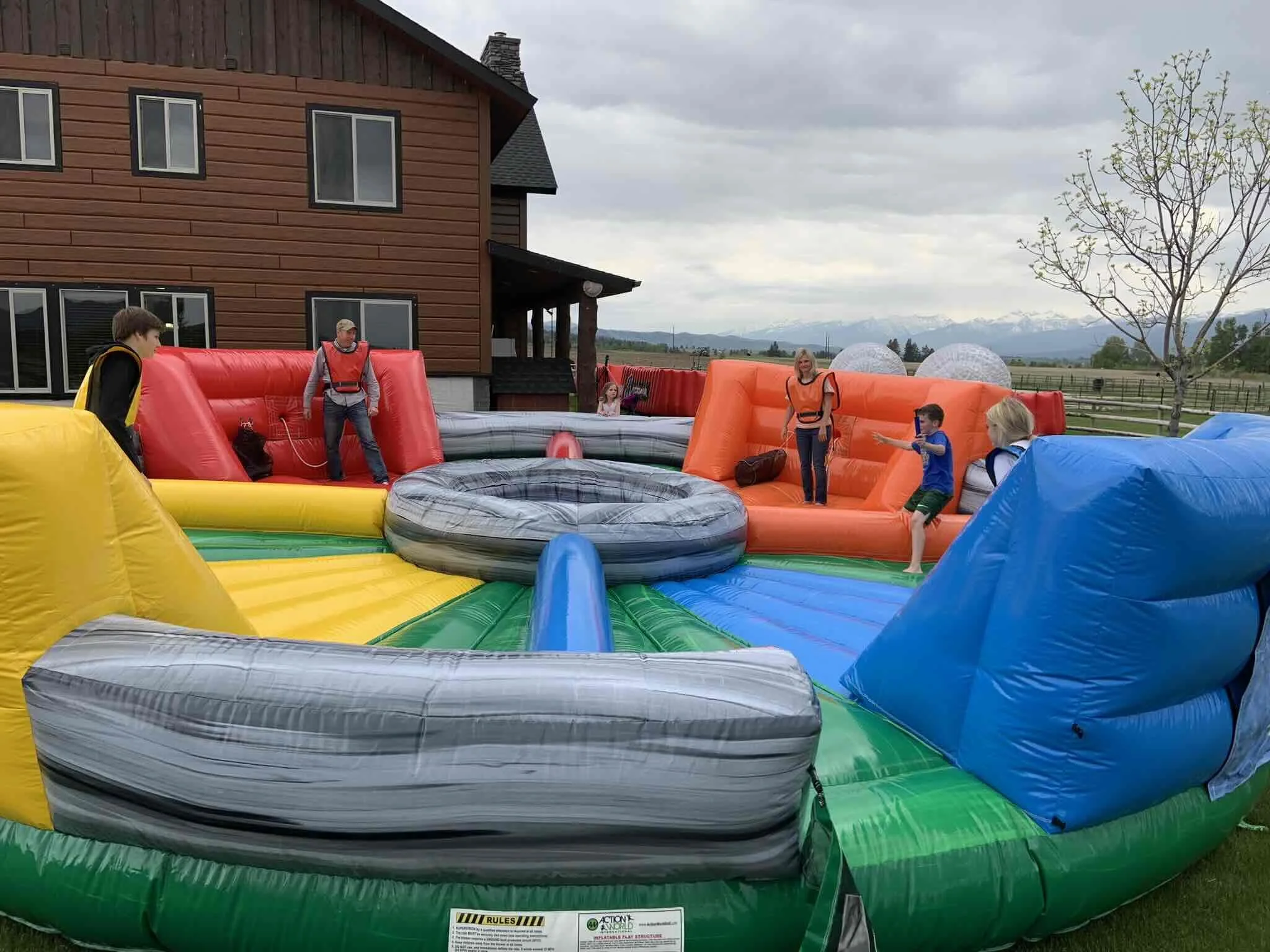 Children and adults playing on a large, colorful inflatable water bounce house outside in a backyard with a house, trees, and mountains in the distance.