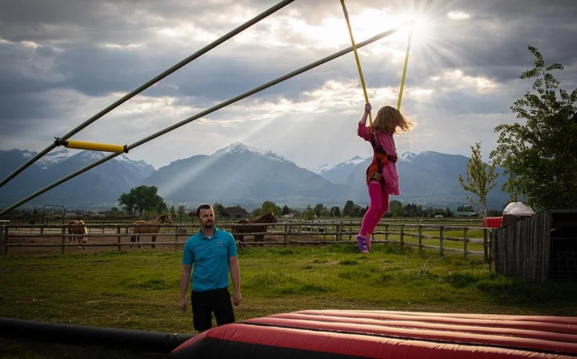A girl in pink and purple swings on a large swing set outdoors with mountains in the background, while a man in a blue shirt looks on. Horses are in a fenced field nearby.