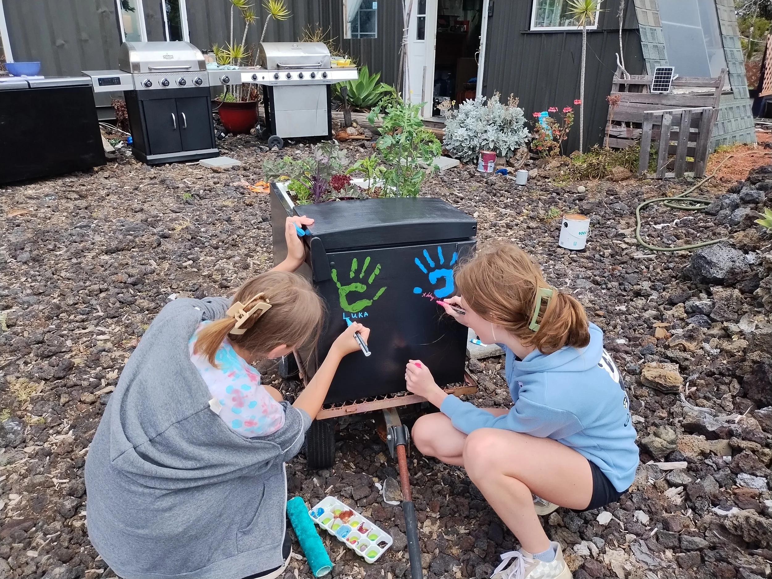 Two young girls are painting colorful handprint designs on a black refrigerator garden in an outdoor backyard with gravel ground, plants, and barbeque grills in the background.