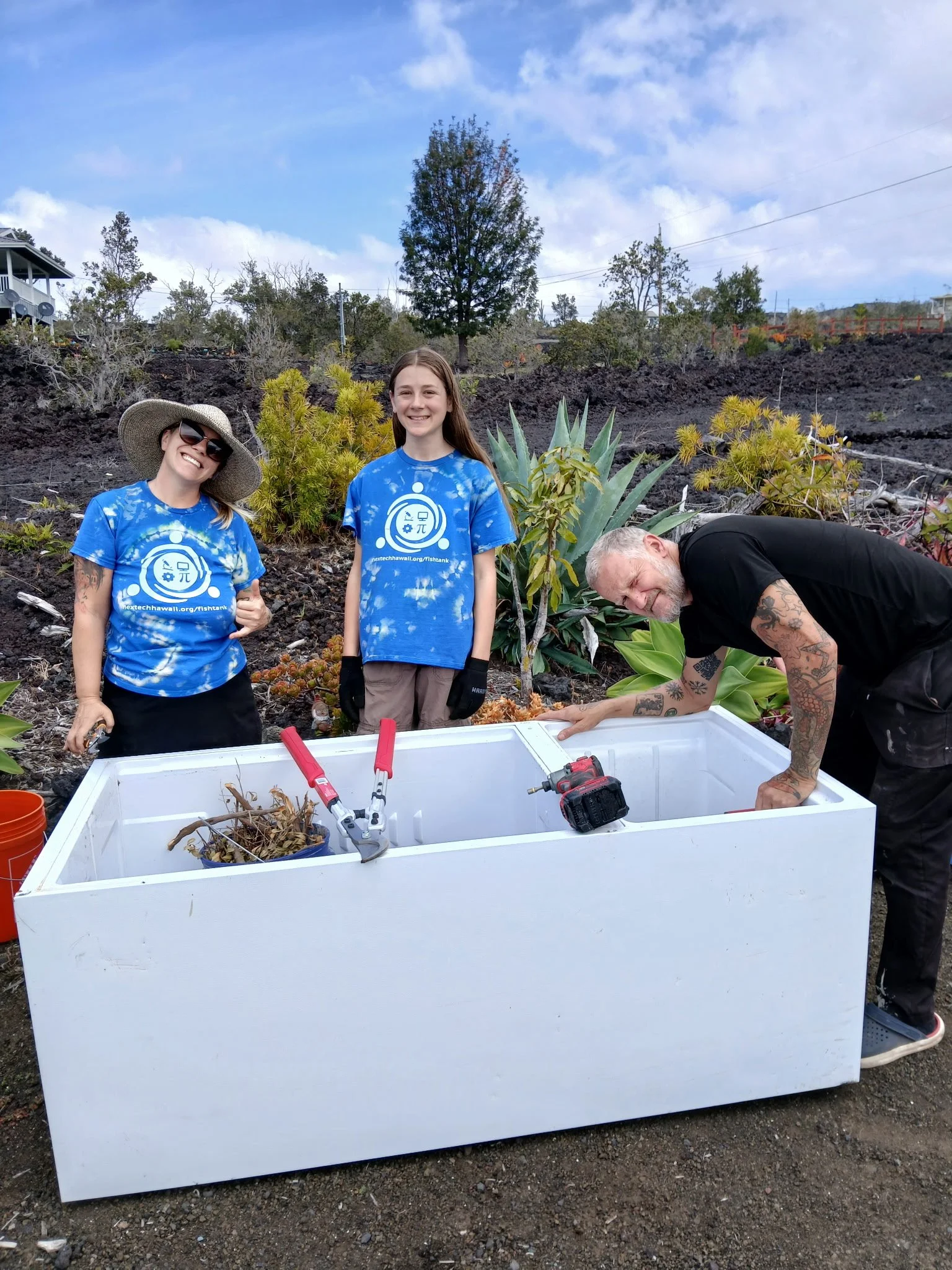 Three people working on a large white recycled refrigerator to turn it into a garden.  Two women and a man are smiling and engaged in gardening or planting activity.
