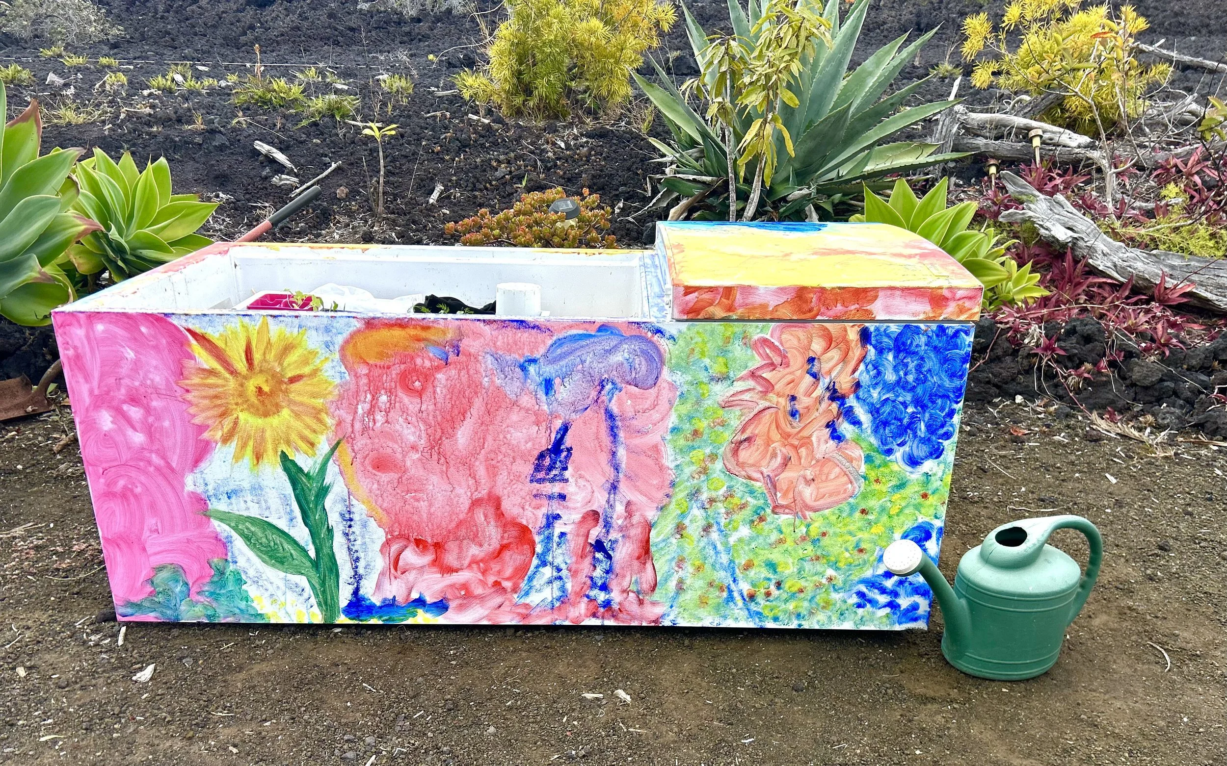 A recycled refrigerator garden decorated with colorful, childlike paintings of flowers and a bright yellow sun, sitting on the ground outdoors with soil and green plants in the background, alongside a green watering can.