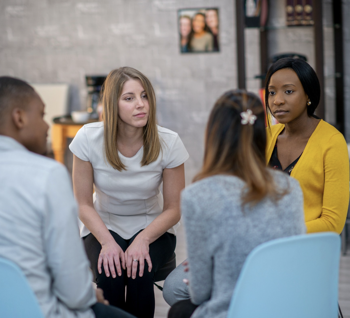 Four women engaged in a serious conversation in a casual setting, with a woman in a white shirt listening attentively and a woman in a yellow cardigan speaking.