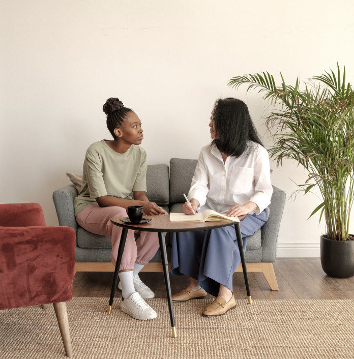 Two women having a conversation in a therapy session, sitting on a gray sofa with a small round table, one taking notes and the other with a cup.