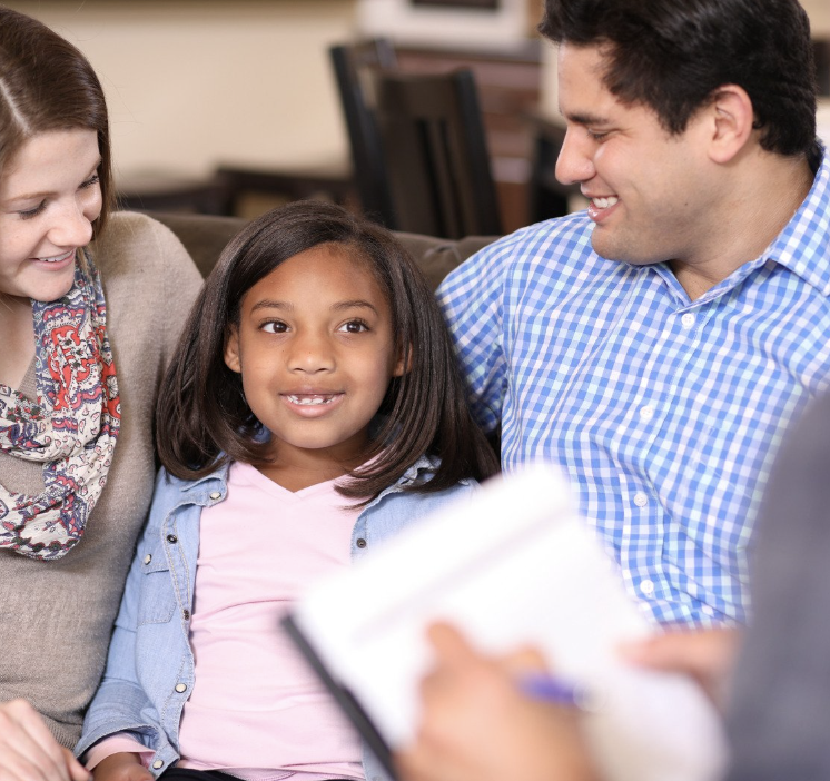 A family of three sitting on a sofa with a therapist in an office. The young girl is smiling while her parents look at her lovingly. The therapist is taking notes.