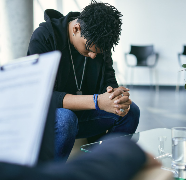 A young man with dreadlocks and a black hoodie sitting with head bowed and hands clasped in a contemplative or prayerful pose in a modern room.