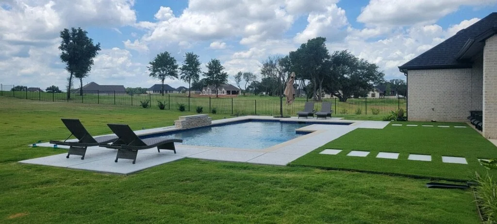 Backyard with a swimming pool, lounge chairs, a patio umbrella, stepping stones on a grassy area, and a house with a brick wall in a suburban neighborhood.