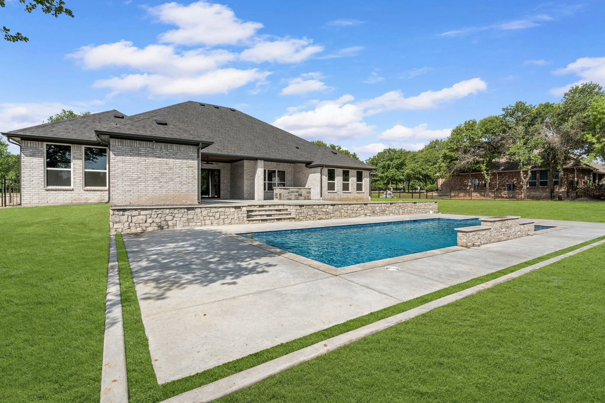 A backyard with a rectangular swimming pool, stone steps, a brick house with a covered patio, and a green lawn under a partly cloudy sky. back yard design. pool builder in dfw. texas. hood county. Granbury. turf grass