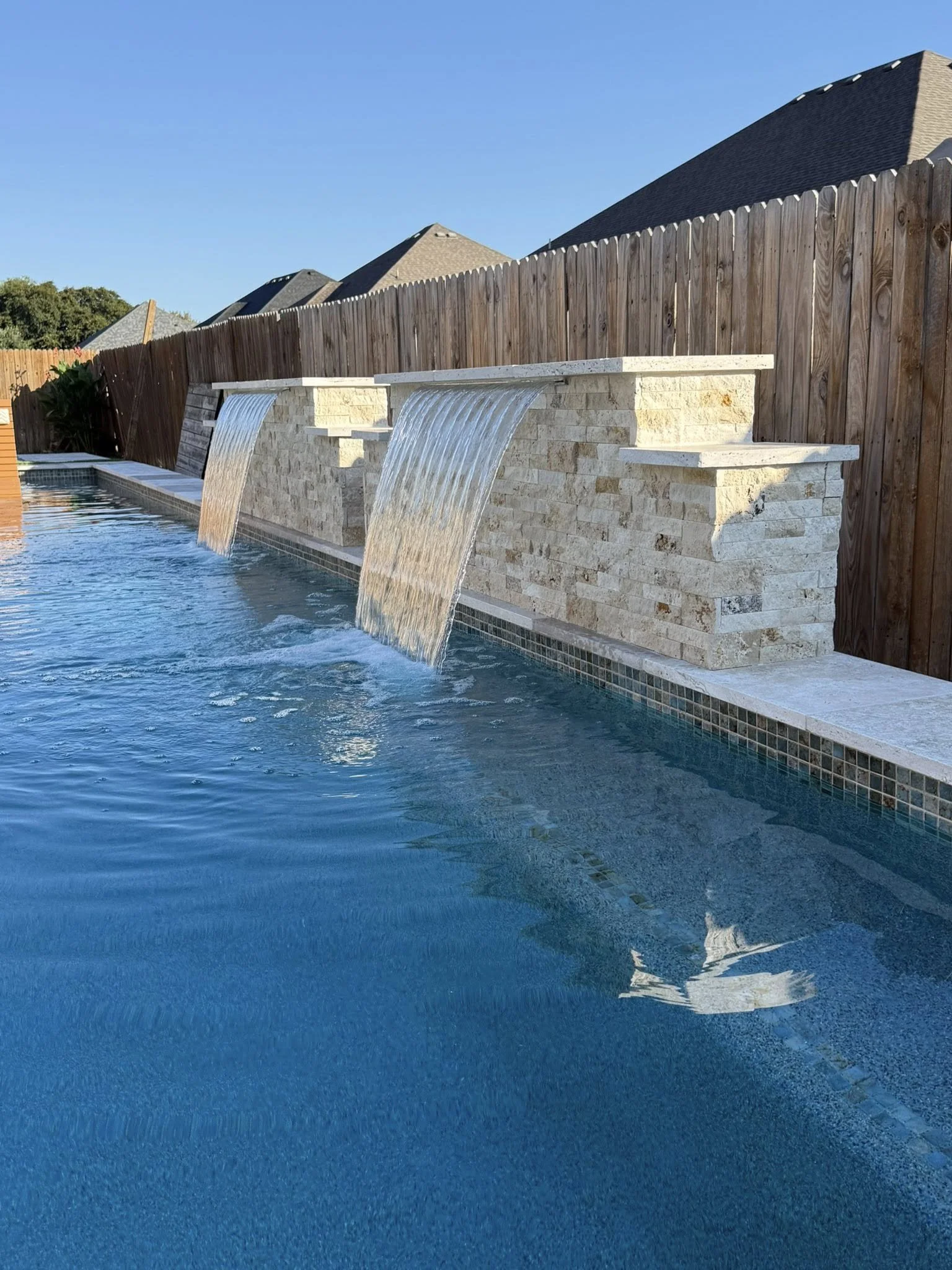 A backyard swimming pool with three small waterfalls cascading over a stone wall with a wooden fence behind it.
