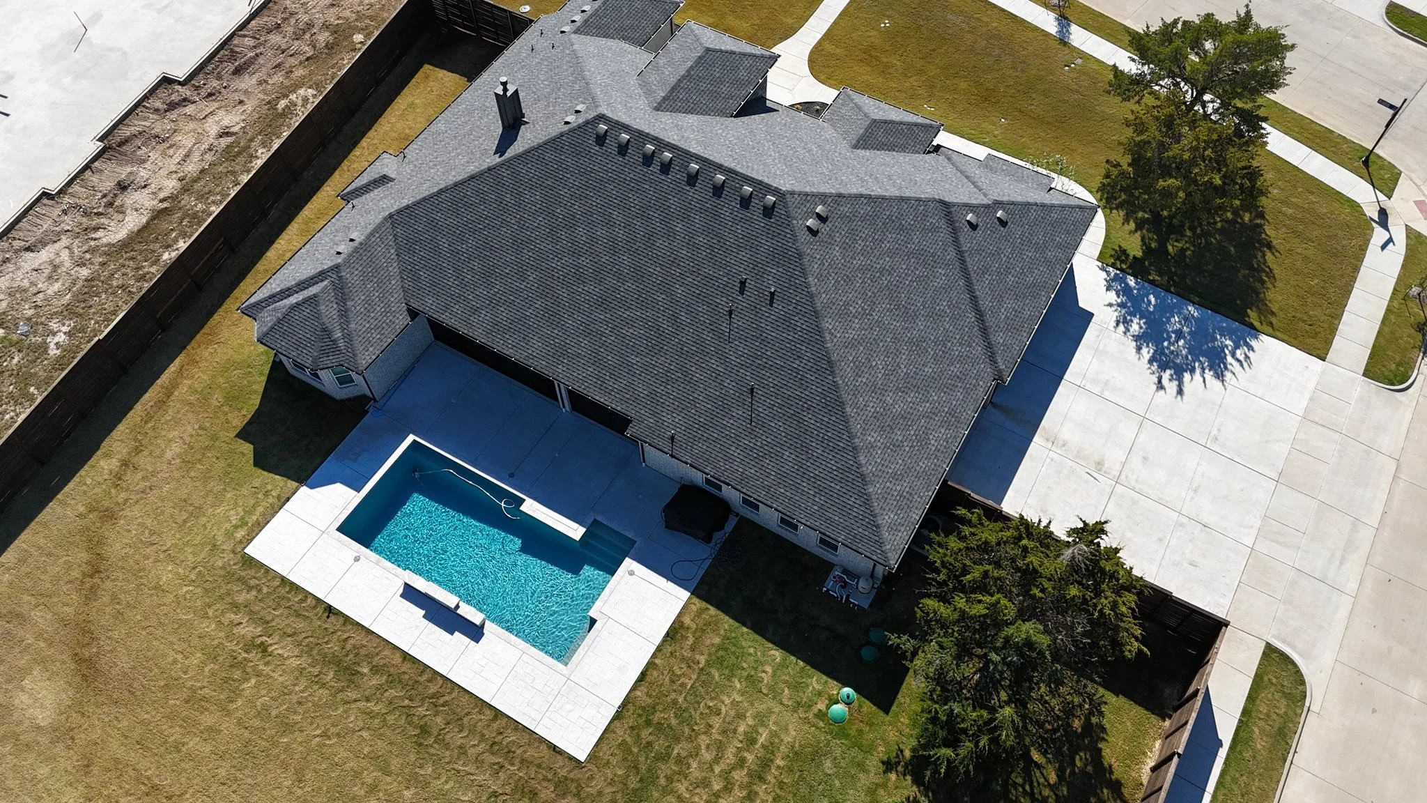 Aerial view of a house with a gray roof, a backyard swimming pool, and a fenced yard. The house is situated in a neighborhood with driveways and green lawns.