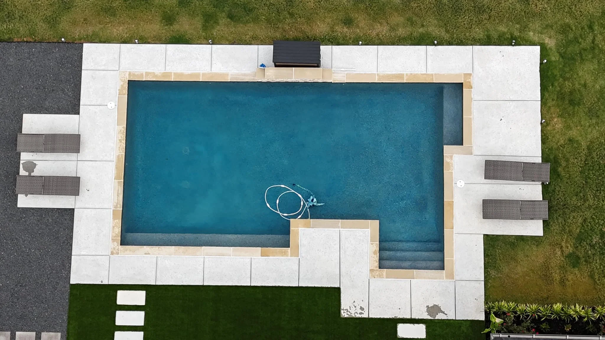 Aerial view of a rectangular swimming pool with a person cleaning it using a pole. The pool is surrounded by concrete paving, with three lounge chairs on each side, and small areas of grass and garden beds around the patio.