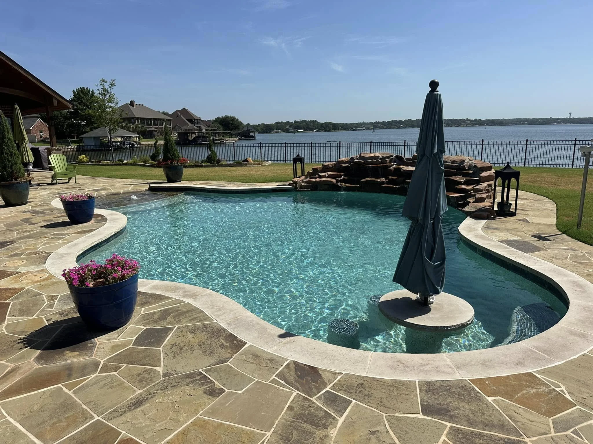 A backyard swimming pool with a view of a granbury lake, surrounded by a stone patio, , and a black metal fence. texas. best of hood county. family owned. quality. luxury. ultimate custom pools. fort worth. texas. honest. trusted. great reviews. Best