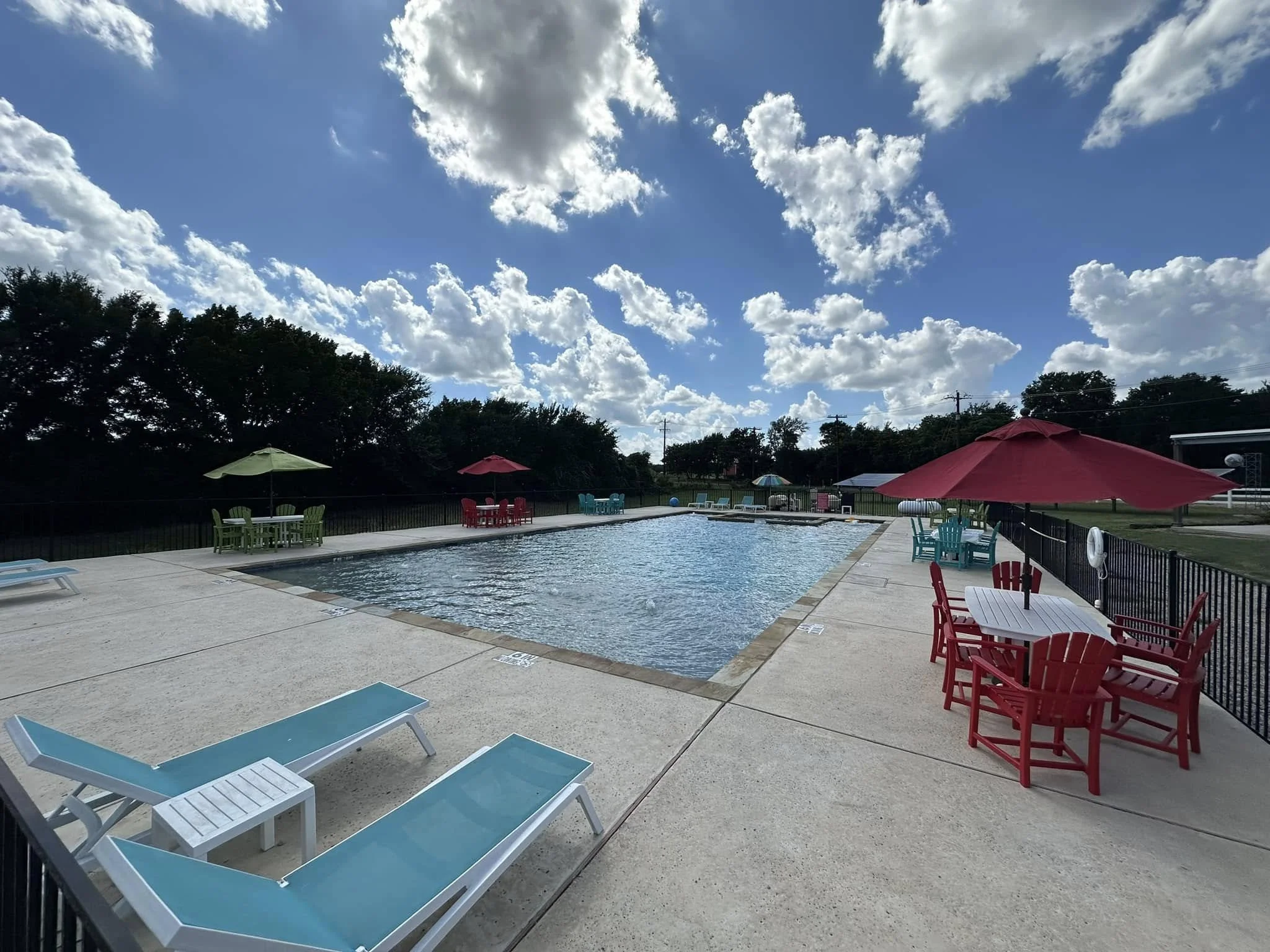 Outdoor swimming pool with colorful tables, chairs, and umbrellas, lounge chairs nearby, set against a backdrop of trees and blue sky with clouds.best of. custom pools.  texas. best of hood county. family owned. quality. luxury. ultimate custom pools