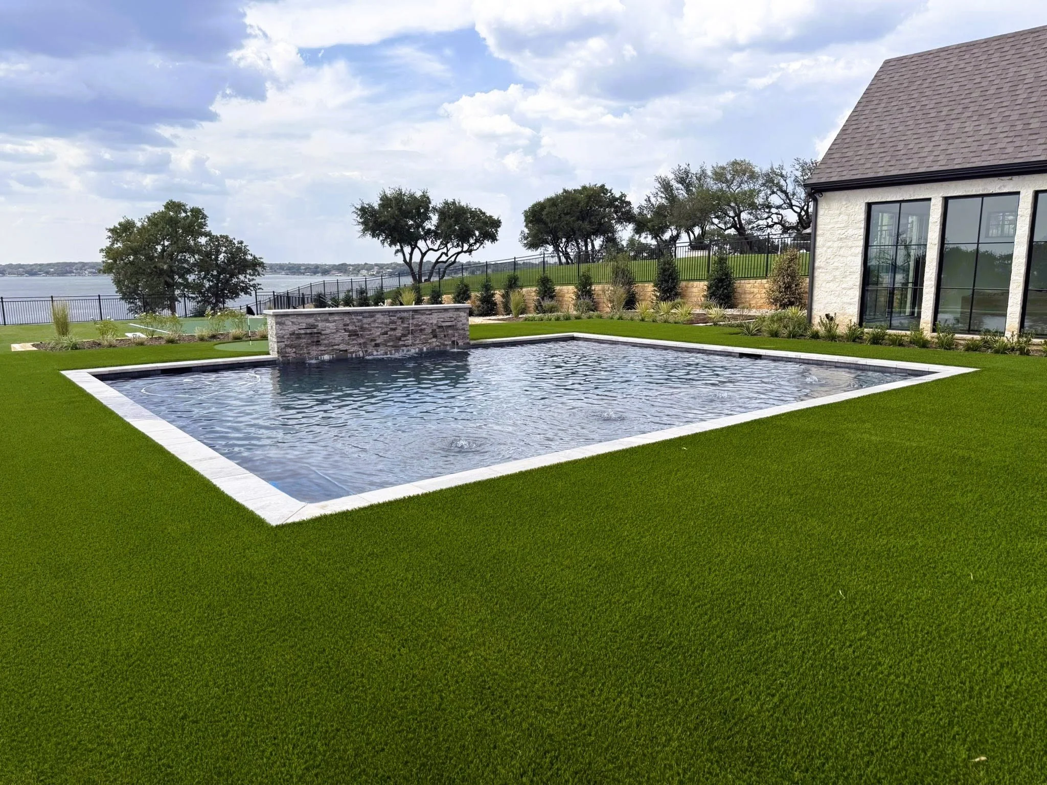 A backyard with a rectangular swimming pool surrounded by a lush green lawn. There is a small waterfall feature on one side of the pool. In the background, there are trees, a lake, and a fence, with a house to the right.