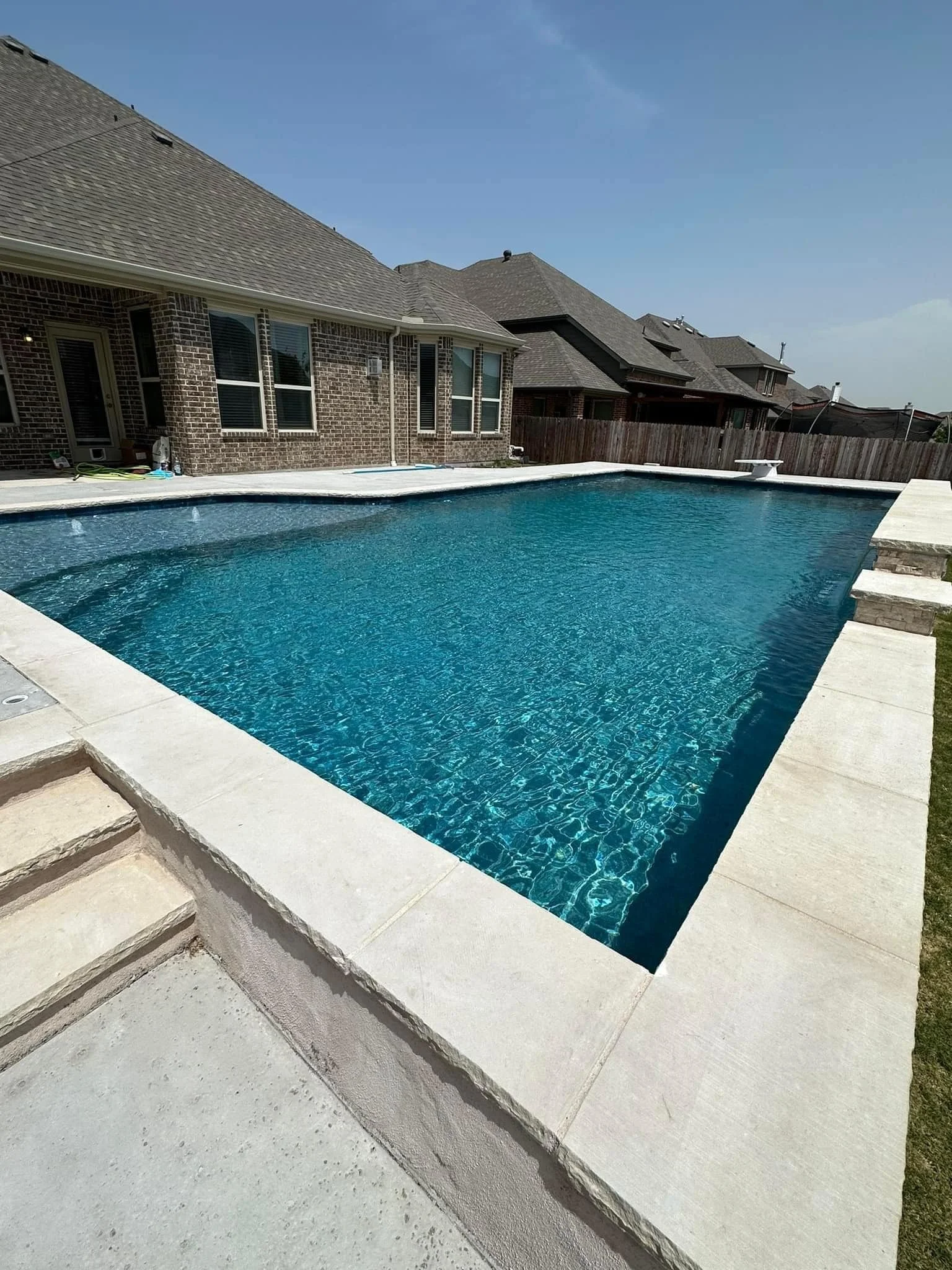 Residential backyard with a rectangular swimming pool surrounded by a concrete deck, a brick house with multiple windows and a sloped roof in the background, clear blue sky.