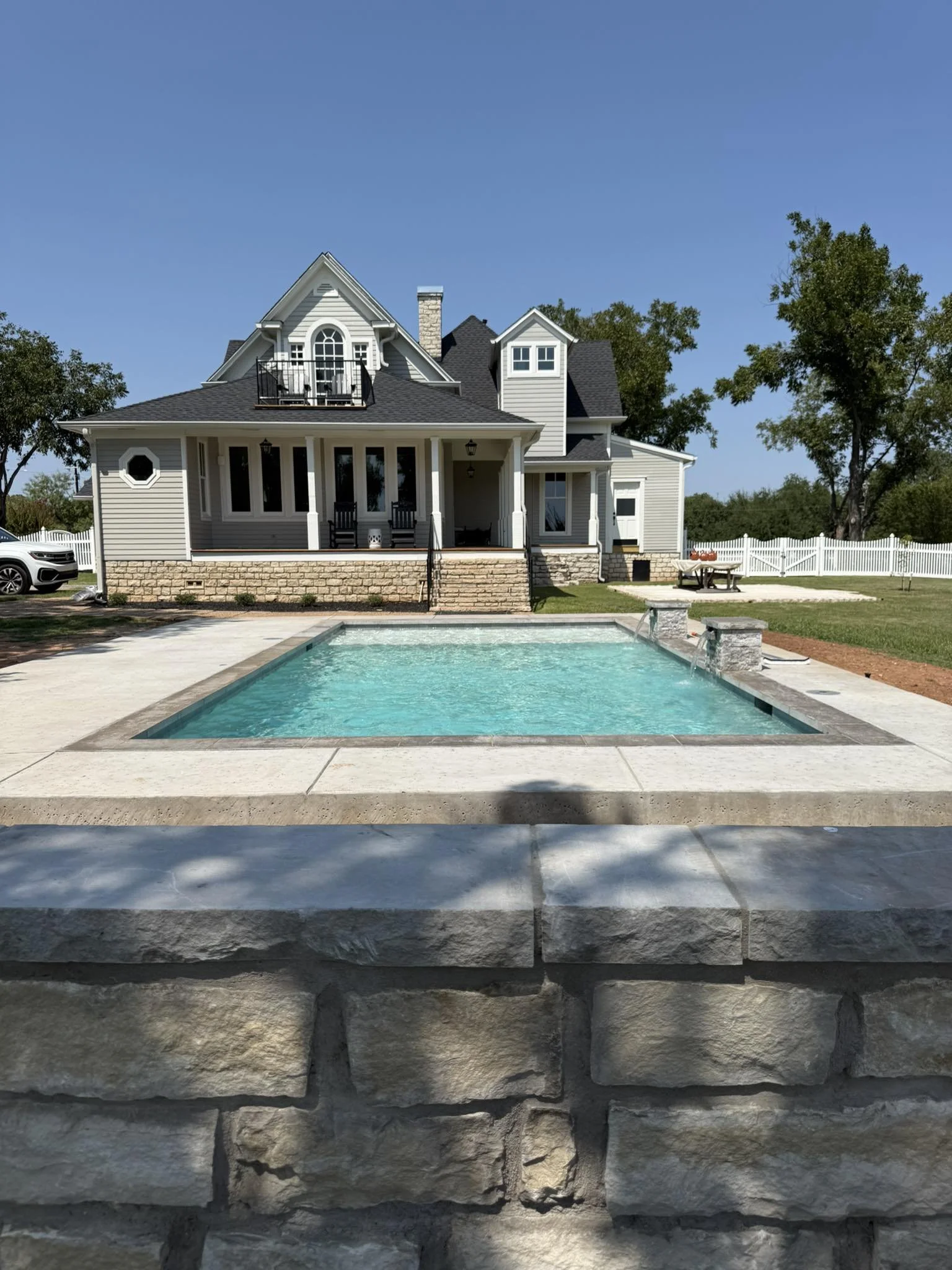 A large house with a front porch, multiple gabled roofs, and white siding, overlooking a swimming pool surrounded by a concrete patio and a stone wall, with trees in the background.