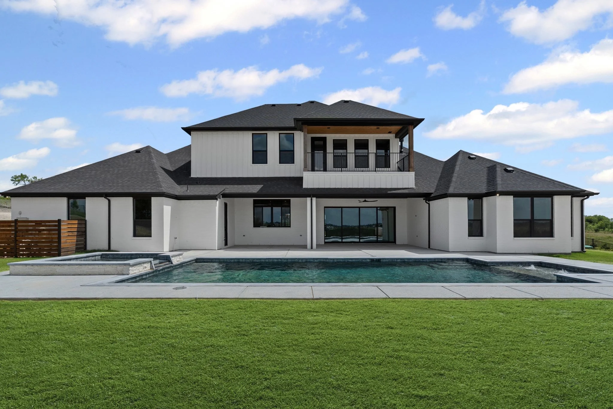 Modern two-story house with a backyard pool, outdoor patio, and green lawn, under a partly cloudy sky.  Custom pool design. Ultimate custom pools. Trusted pool builder.