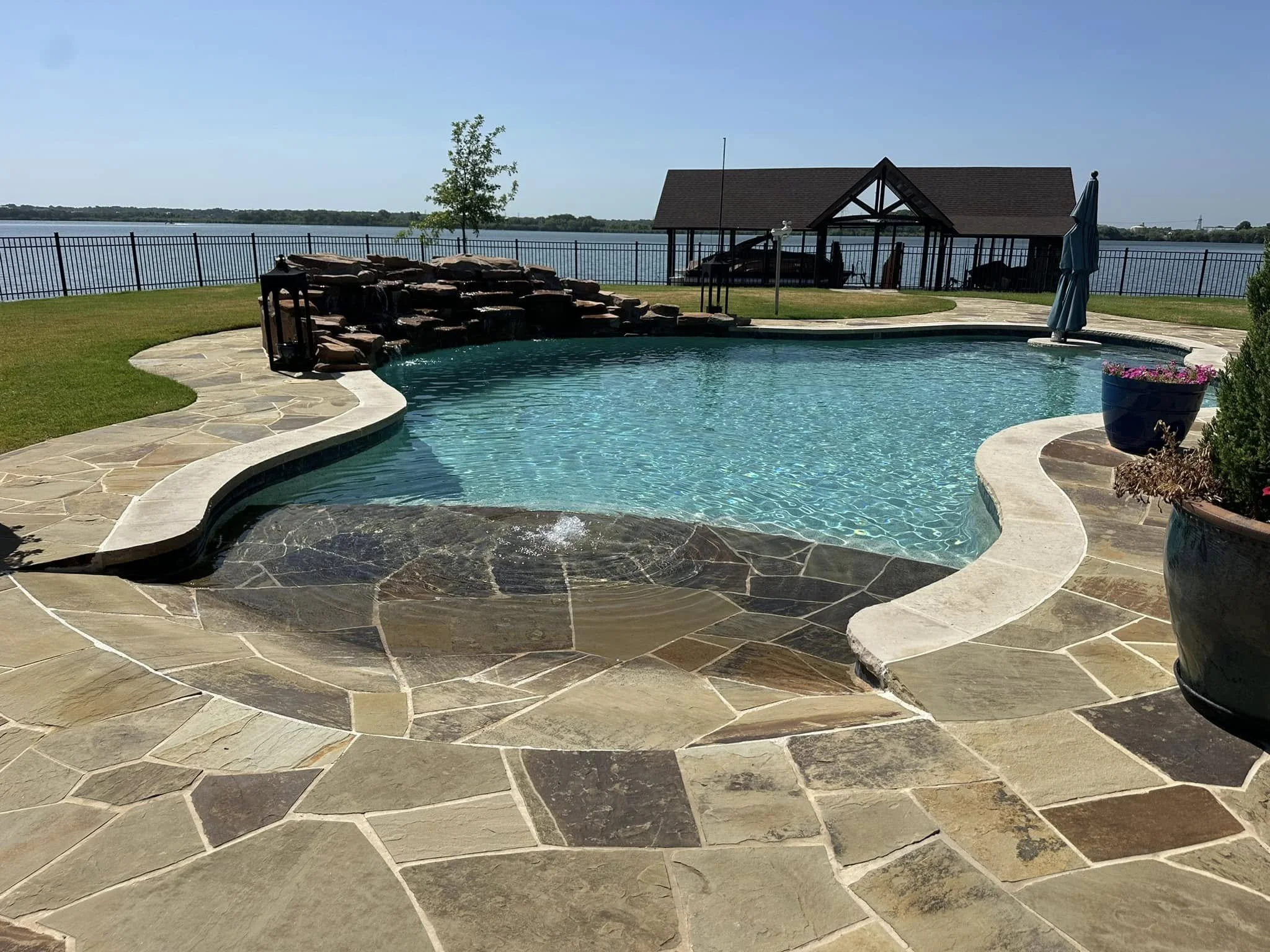 A backyard swimming pool with a waterfall feature, surrounded by a stone deck, overlooking a lake with a fence, and a wooden pavilion in the background.