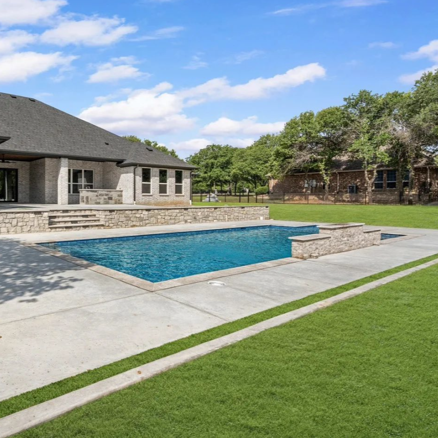 A backyard with a swimming pool, concrete patio, and a brick house with a gray roof, surrounded by green grass and trees under a blue sky with scattered clouds. Custom pool design. Ultimate custom pools. Trusted pool builder.