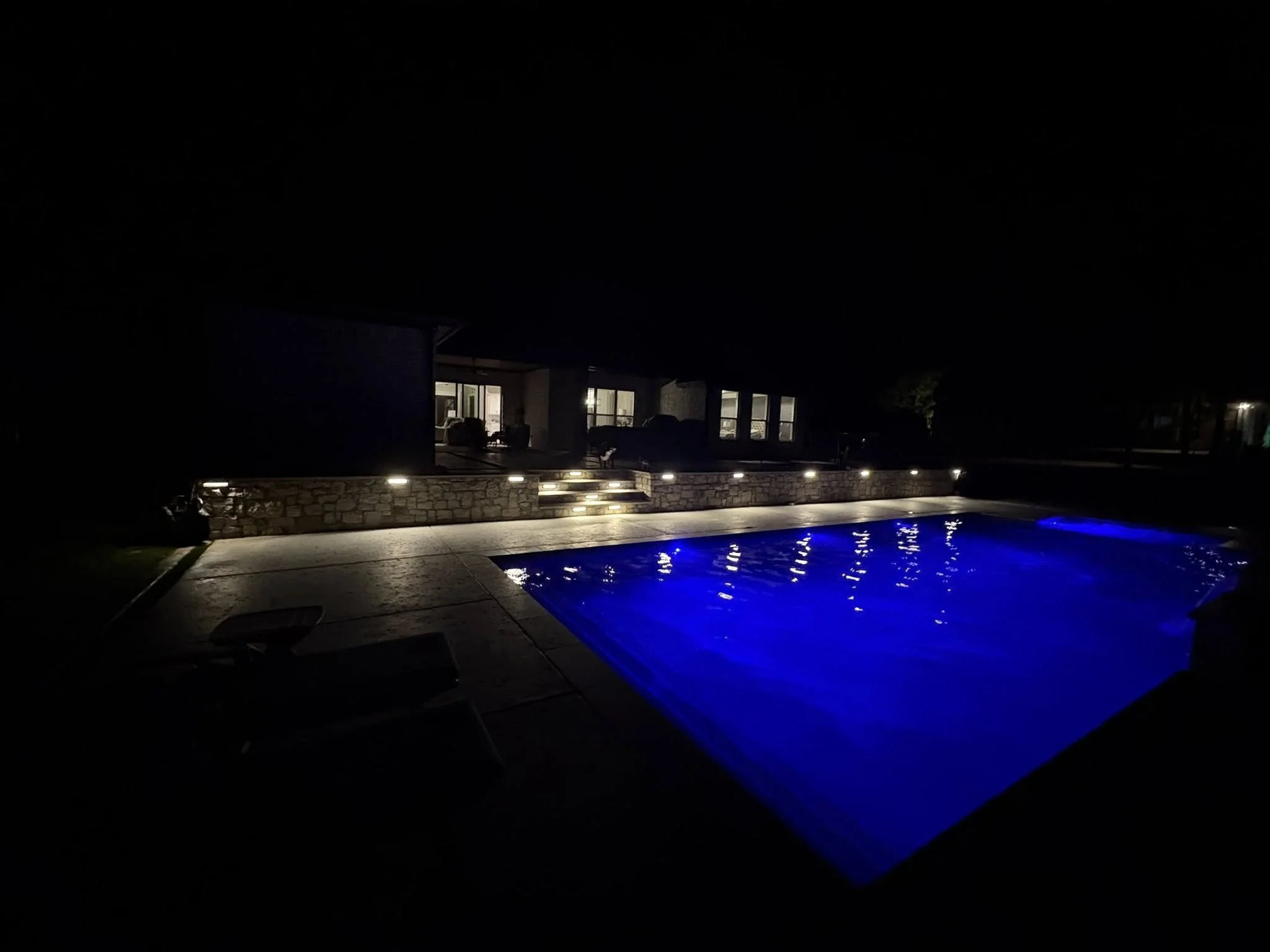 Nighttime view of a backyard pool with blue lighting, surrounded by a concrete deck and a modern house with lit windows.