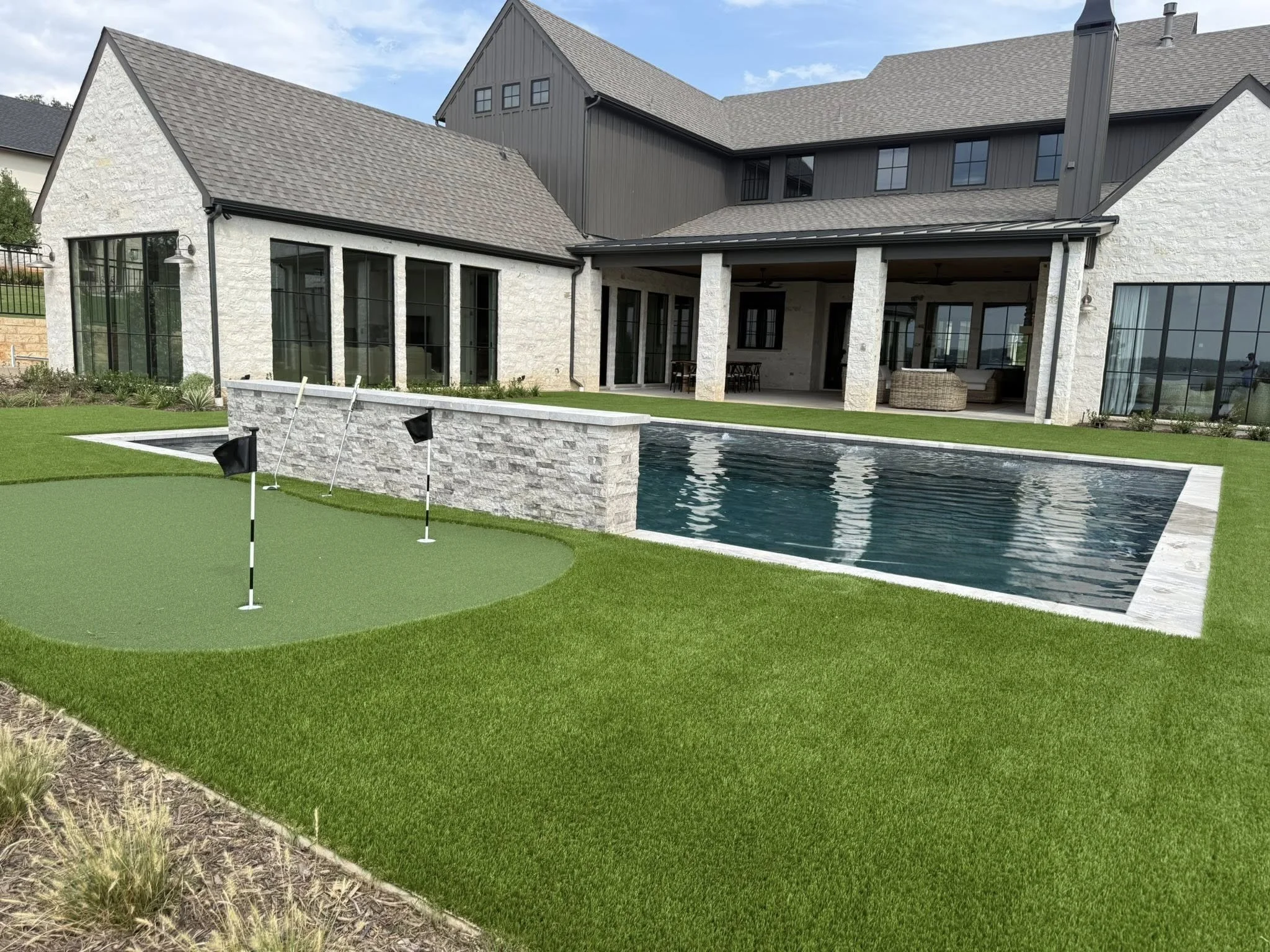 Backyard with artificial grass, a small putting green with three flags, a pool, and a large modern house with covered patio and large windows.