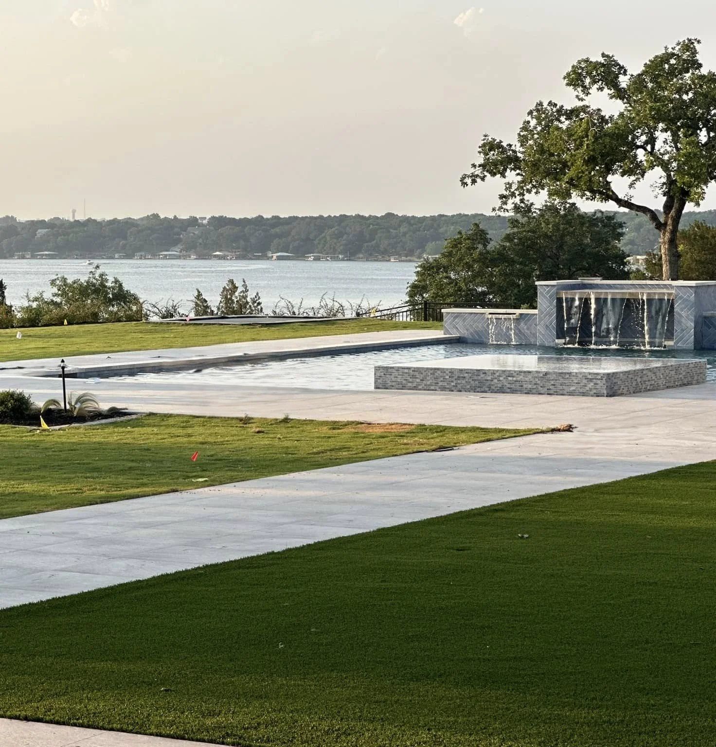 A backyard with a swimming pool near a river or lake, surrounded by green grass, trees, and a paved patio area with a fountain feature.