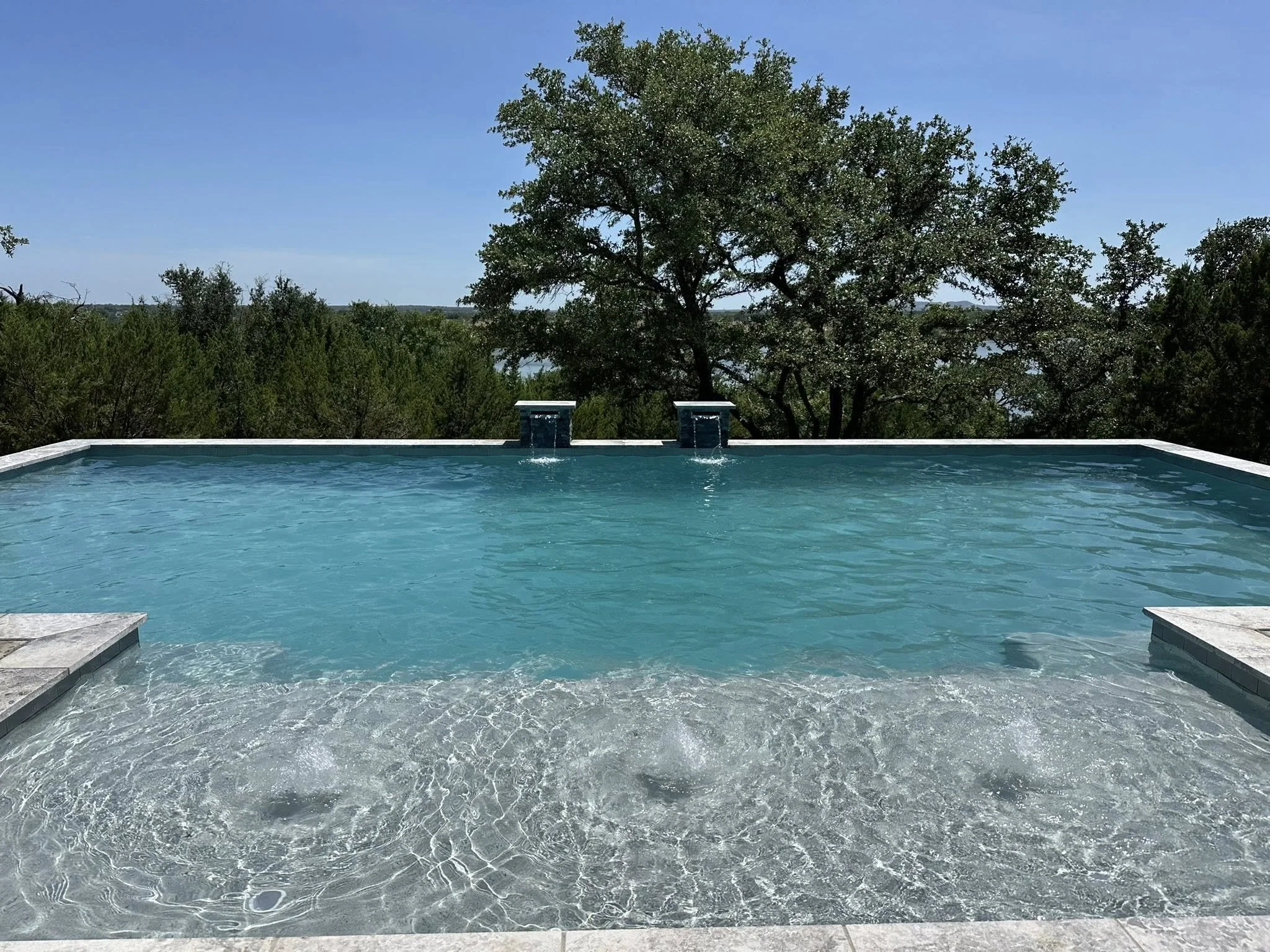 A swimming pool with a clear, blue water surface, surrounded by a stone deck, with two waterfalls at the far end, and trees in the background under a blue sky.