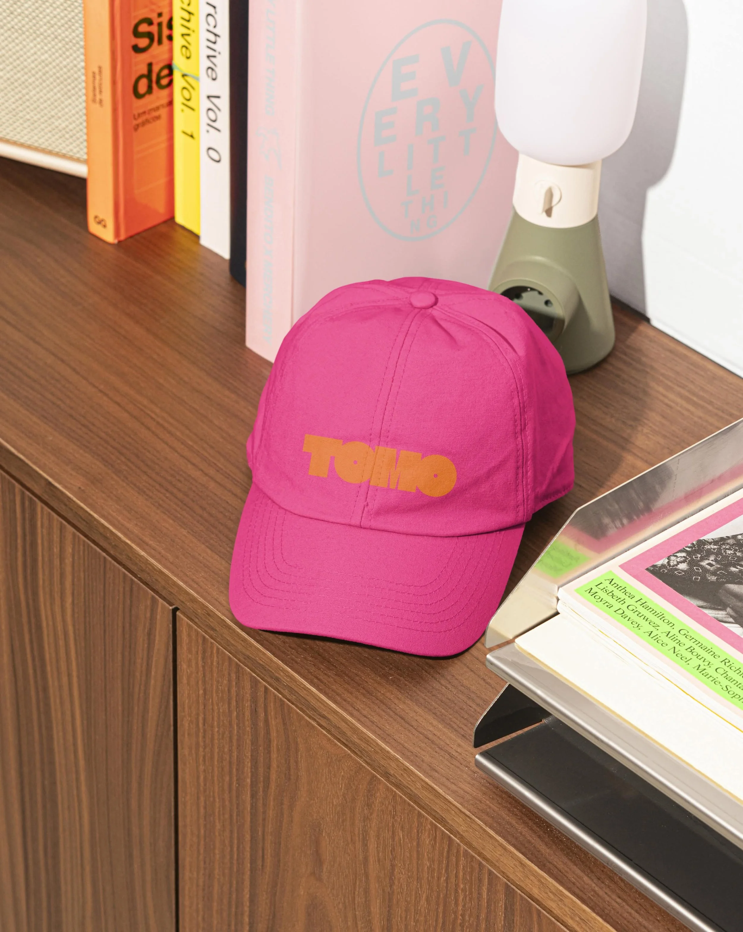 A pink baseball cap with orange and yellow text on the brim, placed on a wooden surface in front of a row of books and a small table lamp.