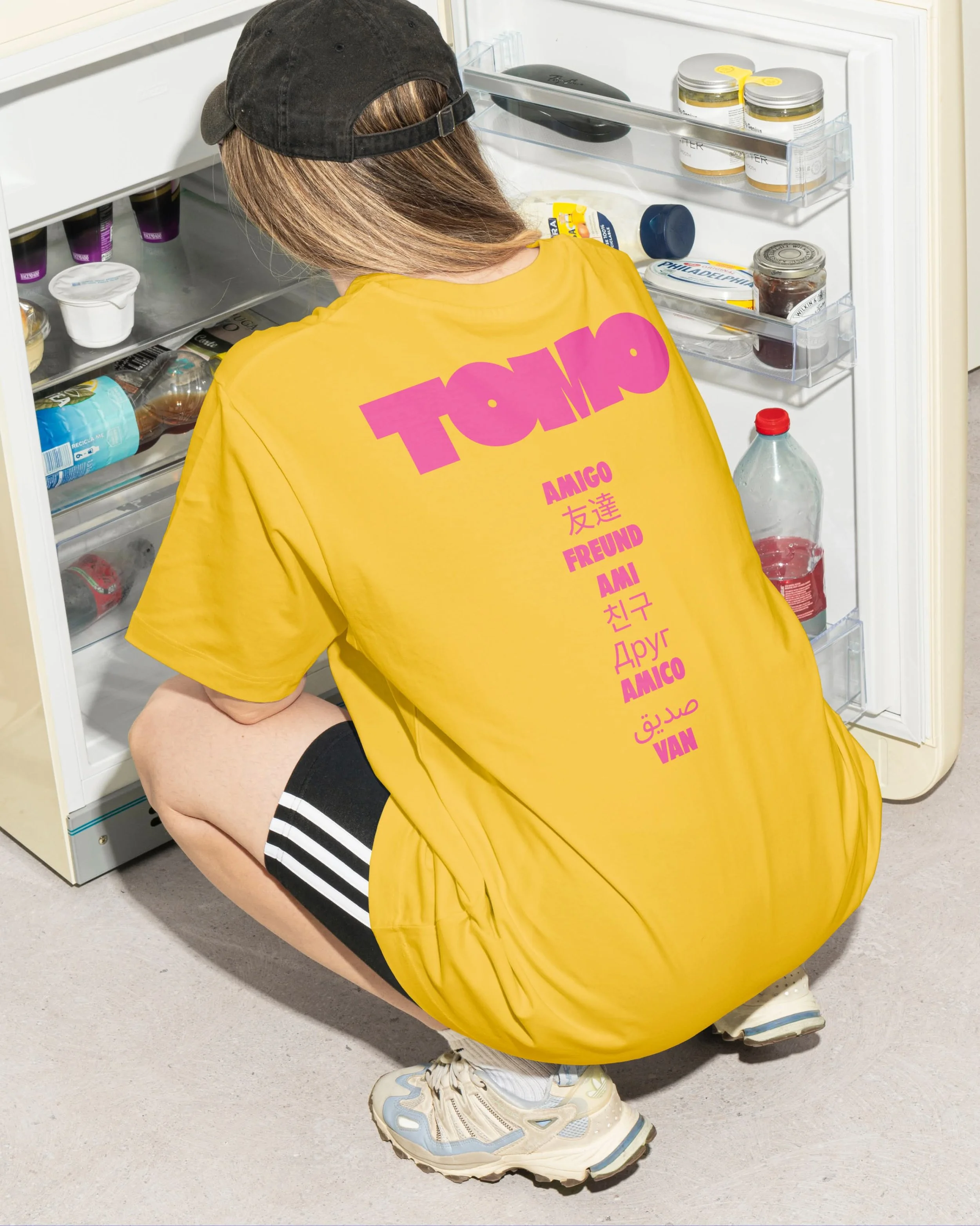 Person wearing a black baseball cap and a yellow T-shirt crouching to look into an open refrigerator with various food items inside. They are wearing black shorts and white sneakers with colorful accents.