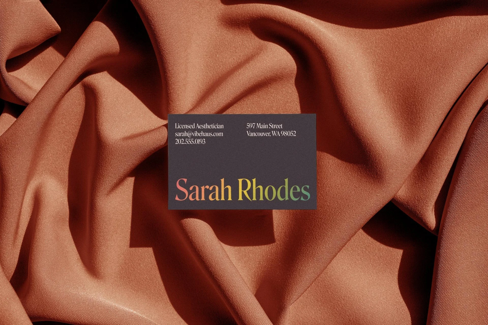 Business card with dark background and colorful text reading 'Sarah Rhodes'. The card is placed on a textured, rippled, peach-colored satin fabric.