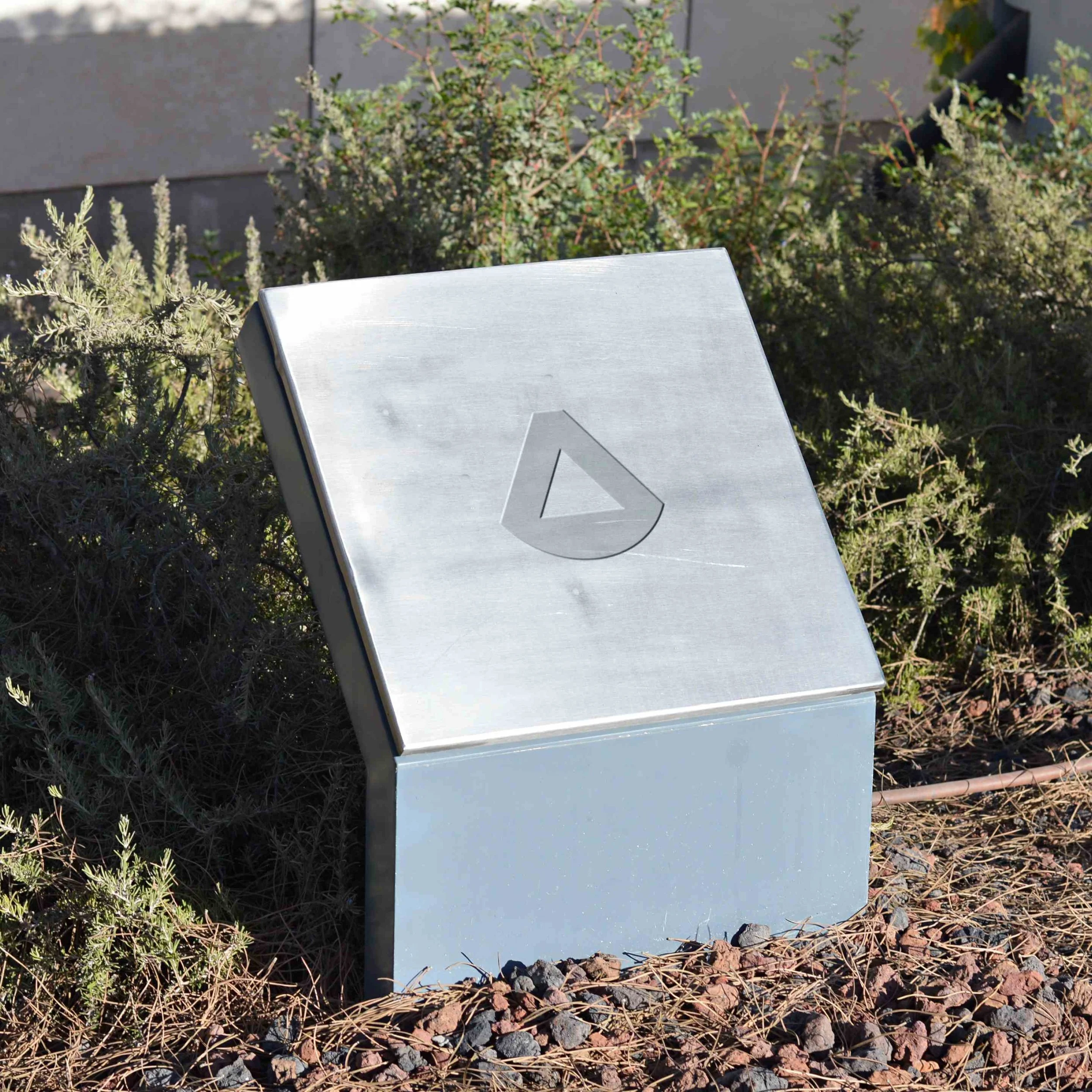 A metallic directional sign with an upward arrow pointing, partially visible, set among bushes and rocks in an outdoor environment.