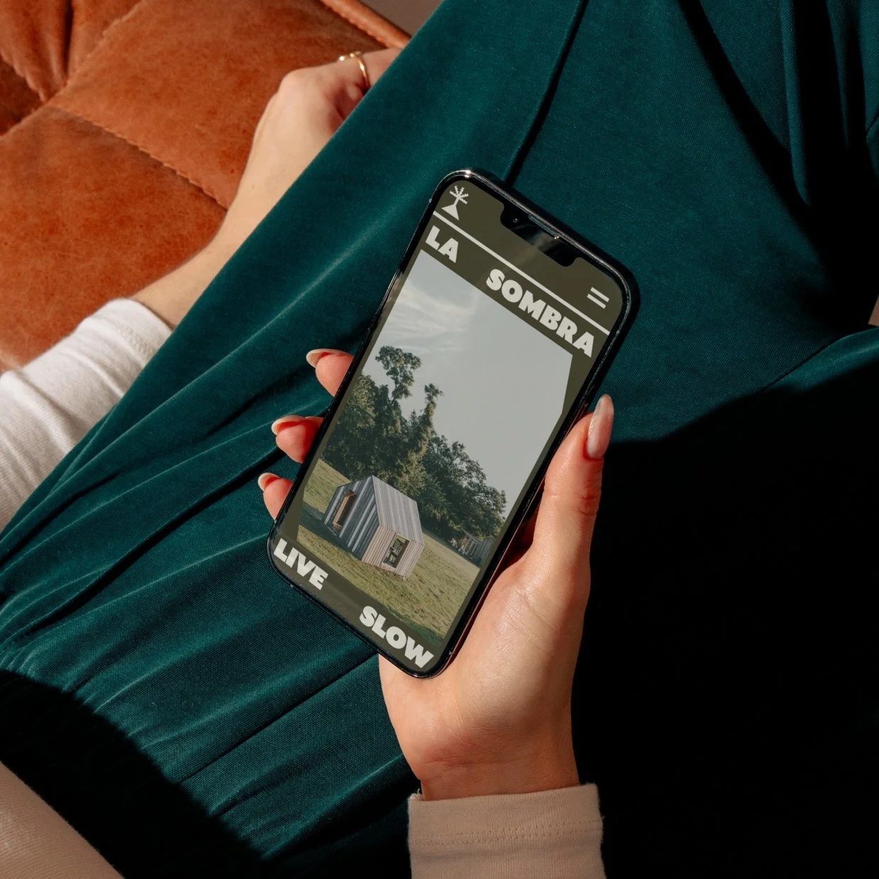 Person holding a smartphone with an app showing a small house with the words 'LA SOMBRA' at the top, and 'LIVE' and 'SLOW' at the bottom. The person is sitting on a green seat with their legs crossed, wearing a cream-colored long-sleeve shirt, and the background has a wooden surface.