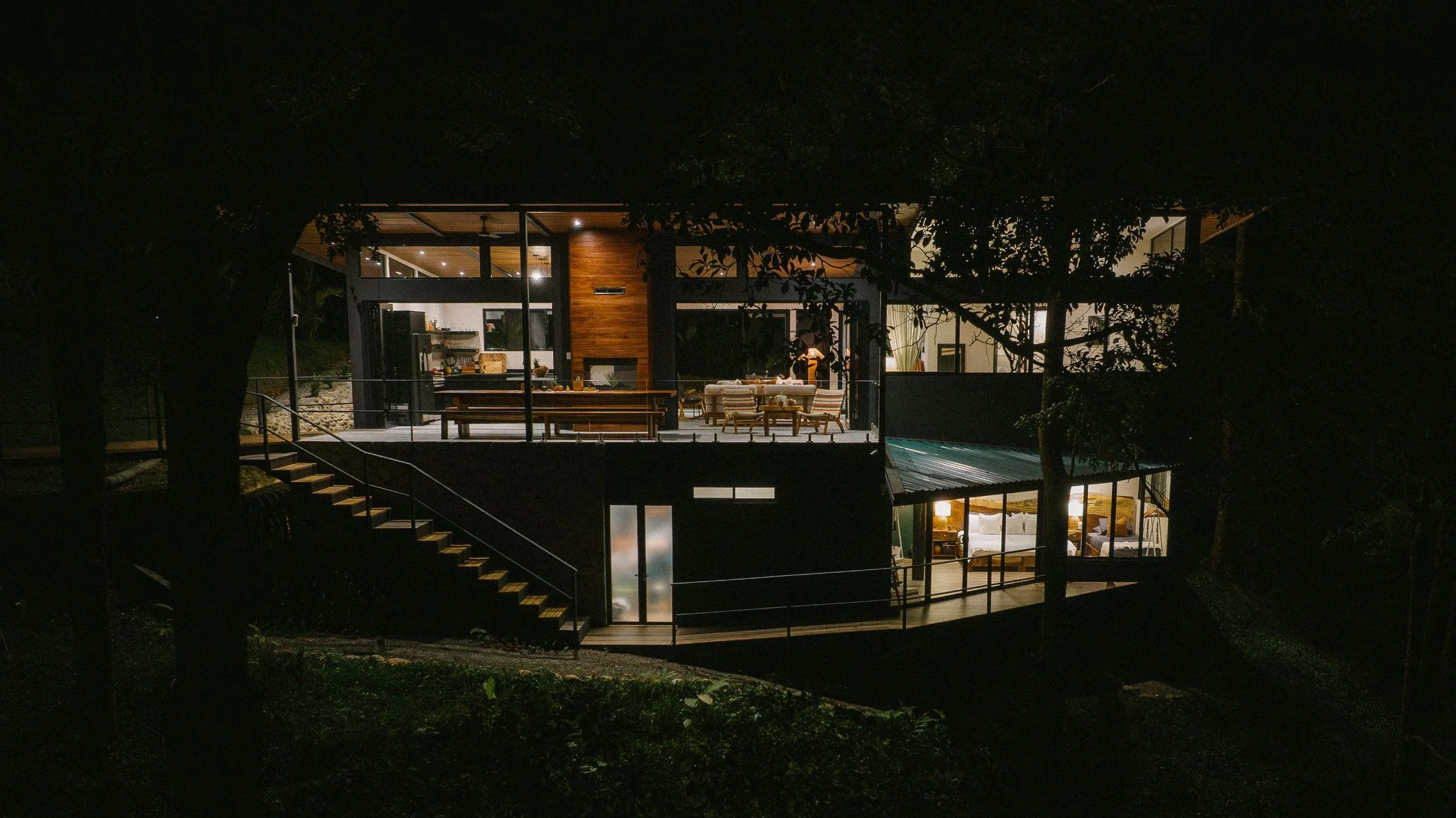 A modern, multi-level house illuminated at night, seen through trees, with outdoor living spaces and large glass windows.