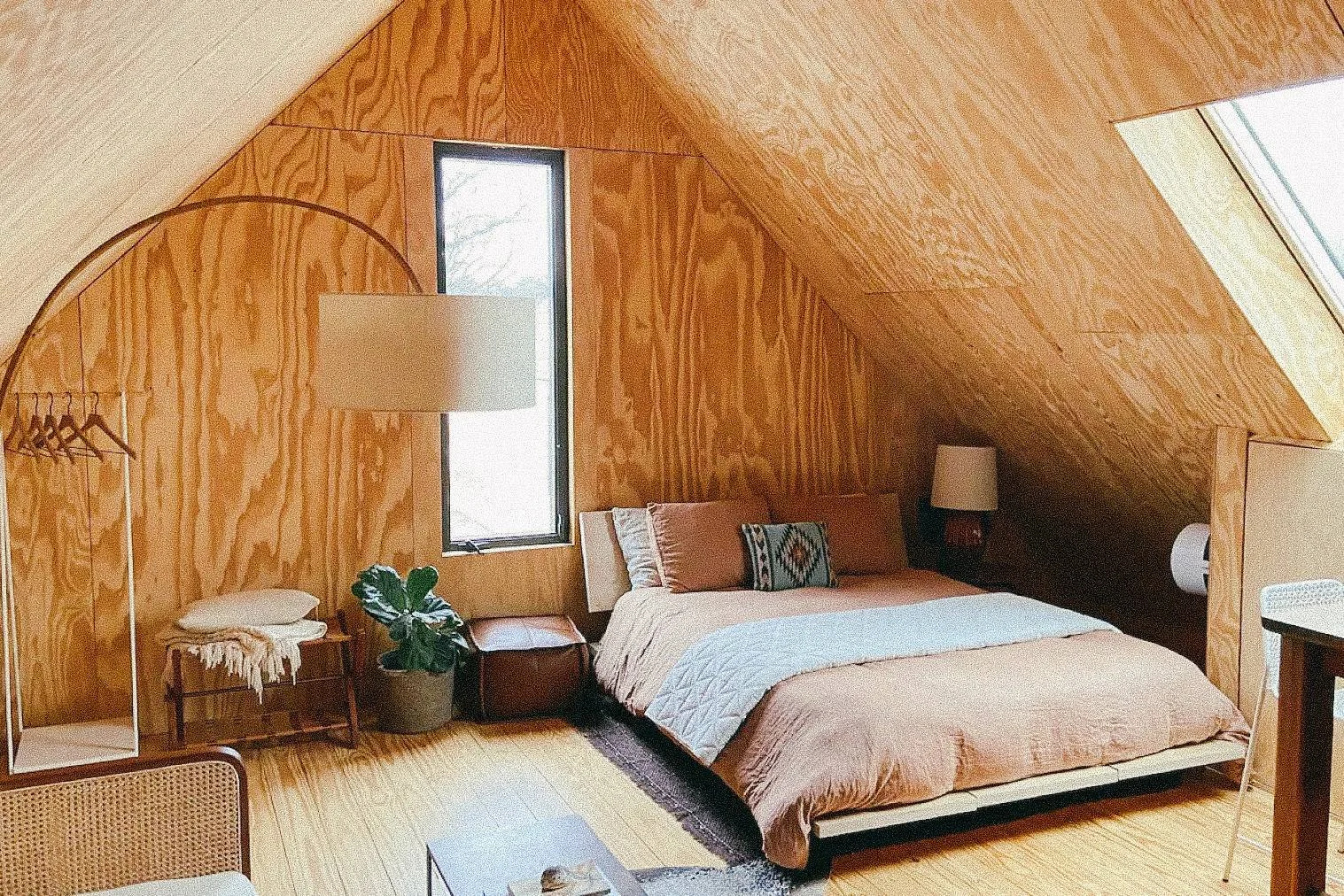 A cozy bedroom with wooden panel walls and sloped ceiling, featuring a bed with beige linens and decorative pillows, a window and skylight, a small side table with a plant, and a standing lamp.