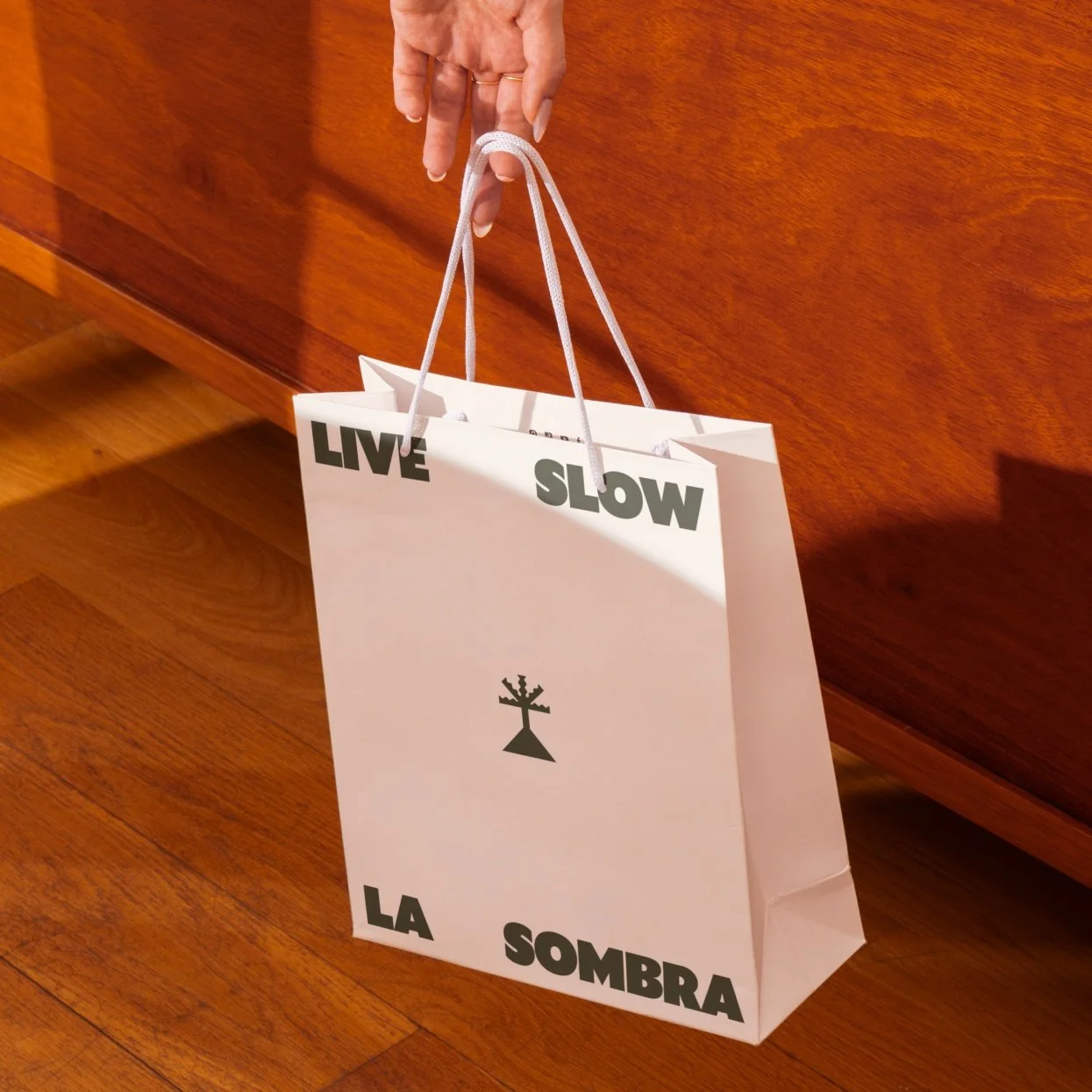 A person is holding a white shopping bag with black text and a logo, standing on a wooden floor against a wooden cabinet. The bag has the words "LIVE SLOW LA SOMBRA" printed on it.