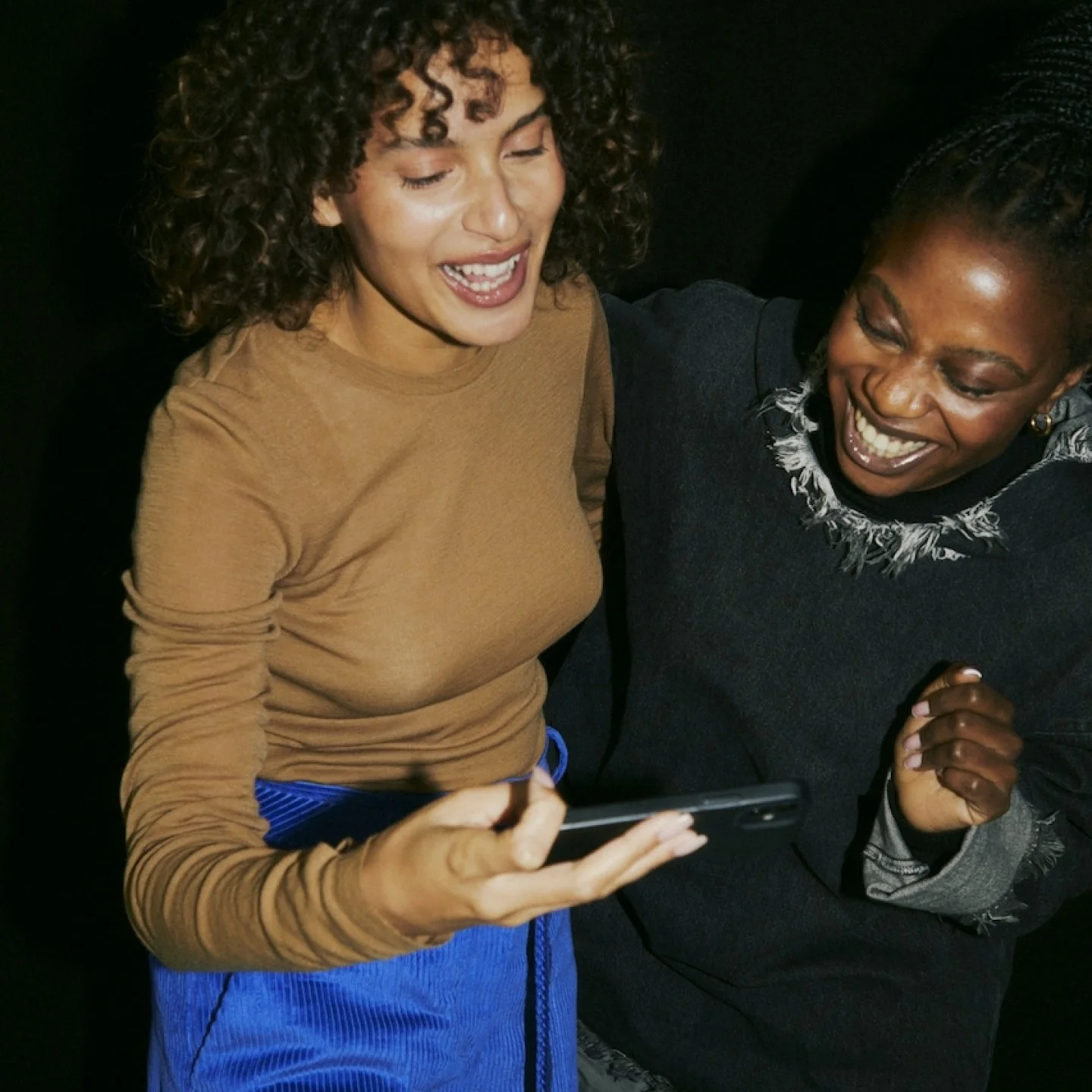 Two women laughing and looking at a phone together, one with curly hair and a tan shirt, the other with braided hair and a black top.