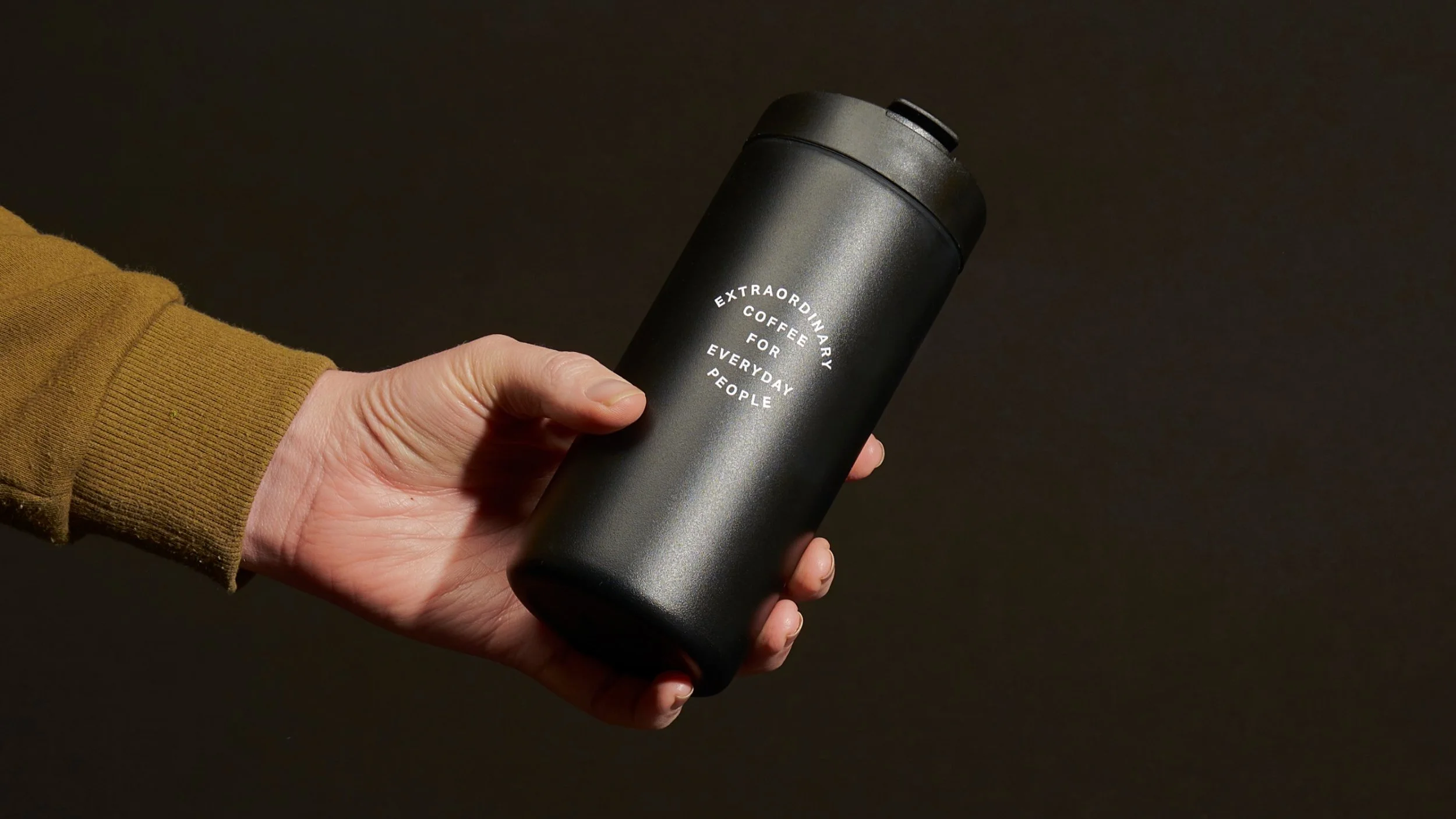 Person holding a black travel mug with white text that reads 'Extraordinary Coffee for Everyday People', wearing a brown sleeve, against a dark background.
