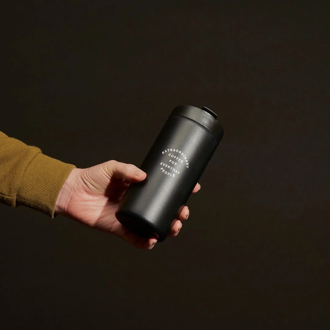 Hand holding a black travel coffee mug with white text that reads 'Extraordinary Coffee for Everyday People' against a dark background.