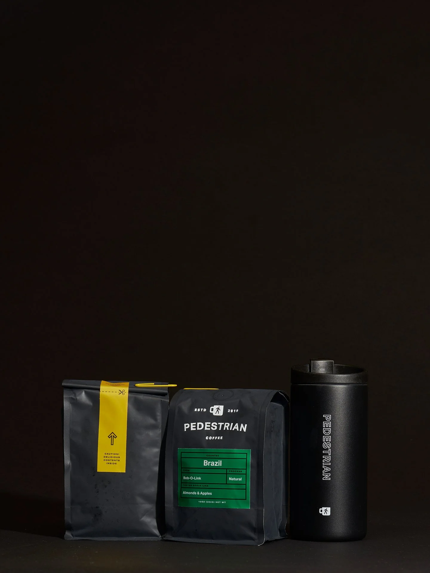 A black bag of Pedestrian coffee with a green label indicating it is Brazil coffee, and a black thermos bottle with white text that reads 'PEDESTRIAN' on a dark background.