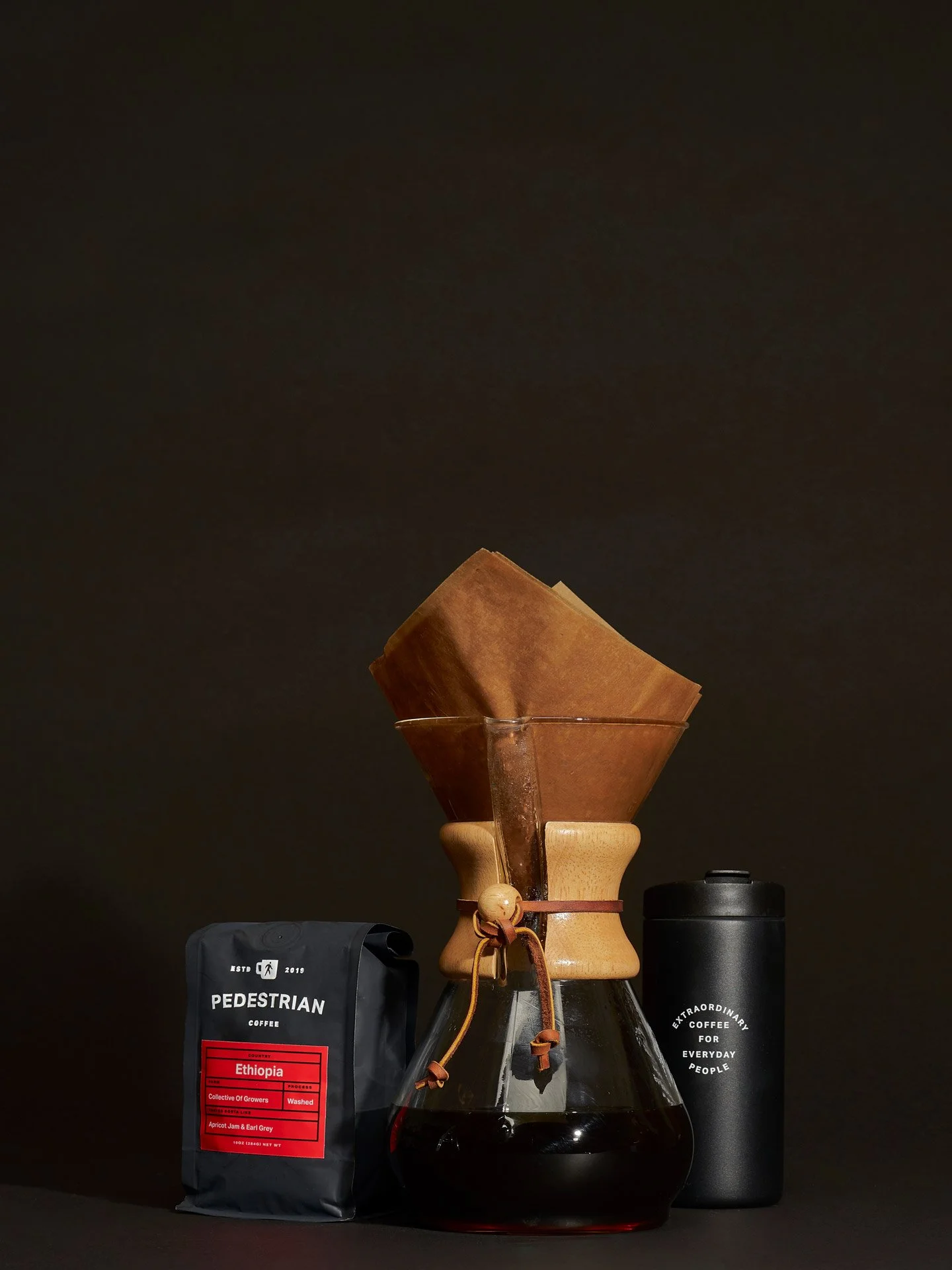 Coffee brewing setup with a Chemex coffee maker, a bag of Ethiopia coffee beans, and a black insulated coffee tumbler against a dark background.