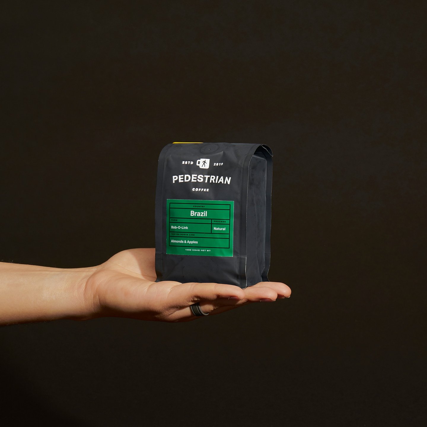 Hand holding a black bag of Pedestrian Coffee labeled with Brazil, natural process, almonds & apples flavor, against a dark background.