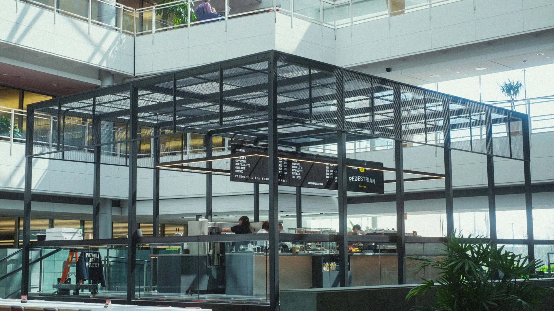 Interior view of a modern shopping mall or airport terminal with multiple levels, escalators, glass walls, and a coffee kiosk in the foreground. There are people sitting at tables and walking around in the background.